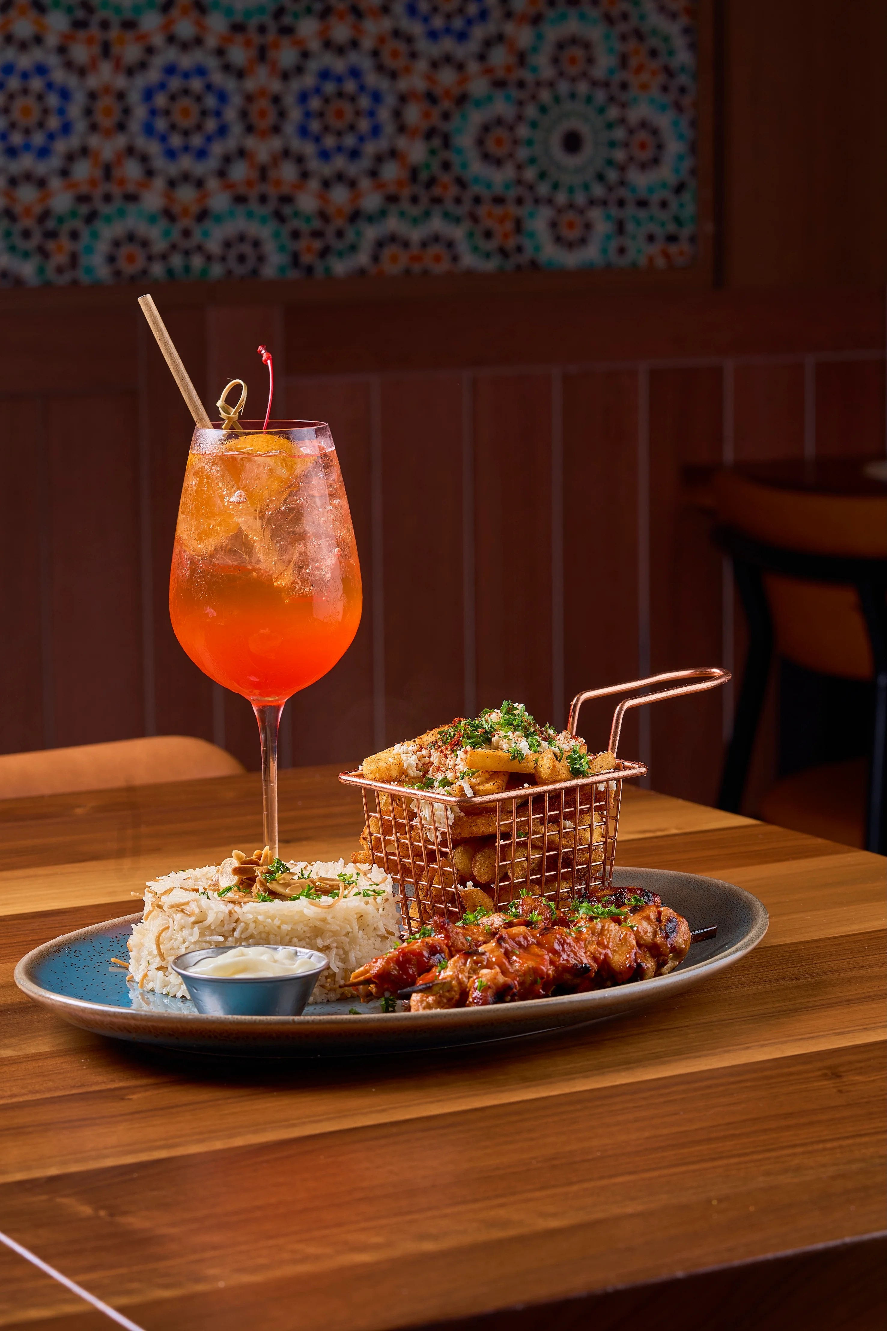 A vibrant meal with grilled skewers, seasoned rice, and fries in a basket, accompanied by an orange cocktail on a wooden table. A colorful mosaic backdrop enhances the warm, inviting ambiance.