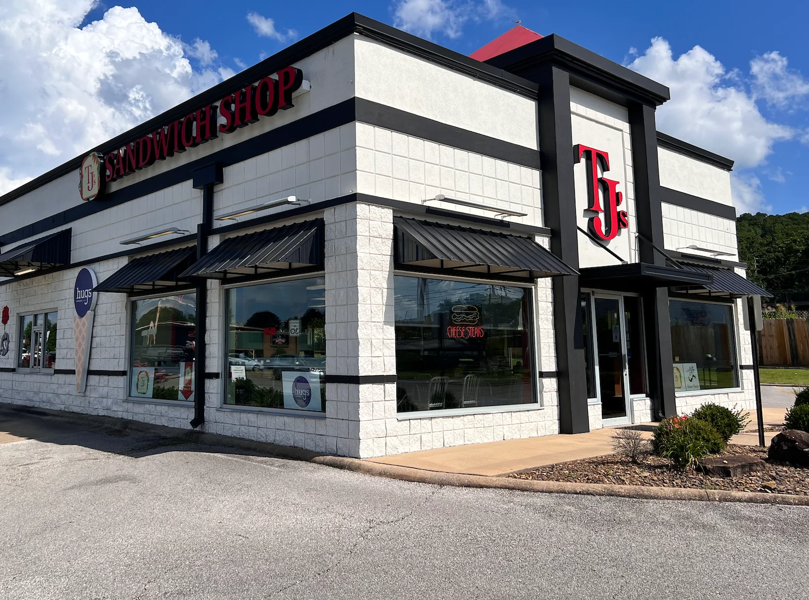 Exterior of TJ's Sandwich Shop with bold signage, mural artwork, and Hugs Ice Cream branding on the windows, under a bright blue sky in a suburban shopping area.