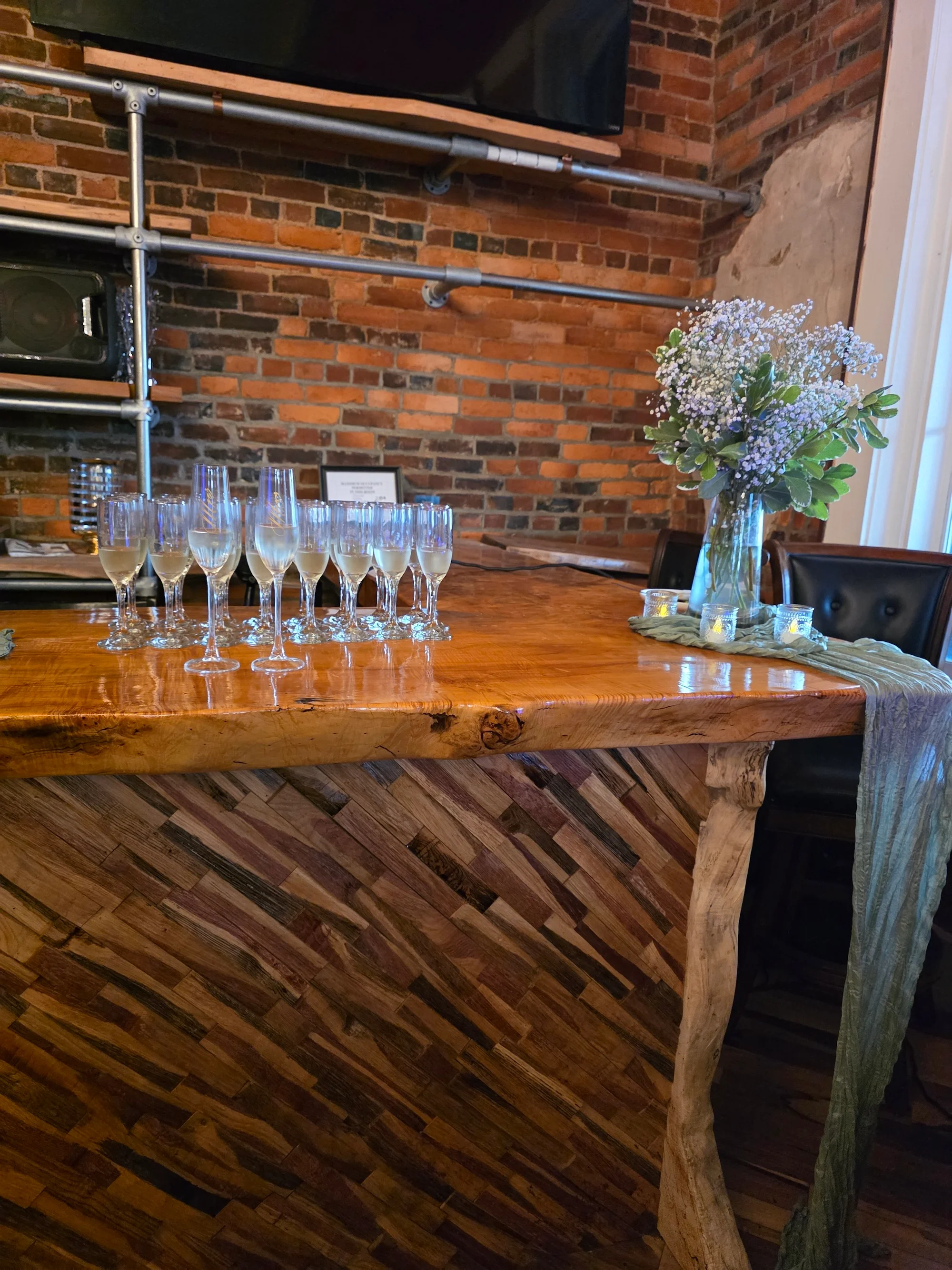 A rustic bar top featuring an arrangement of champagne flutes, a vase with flowers, and a backdrop of exposed brick and industrial decor.