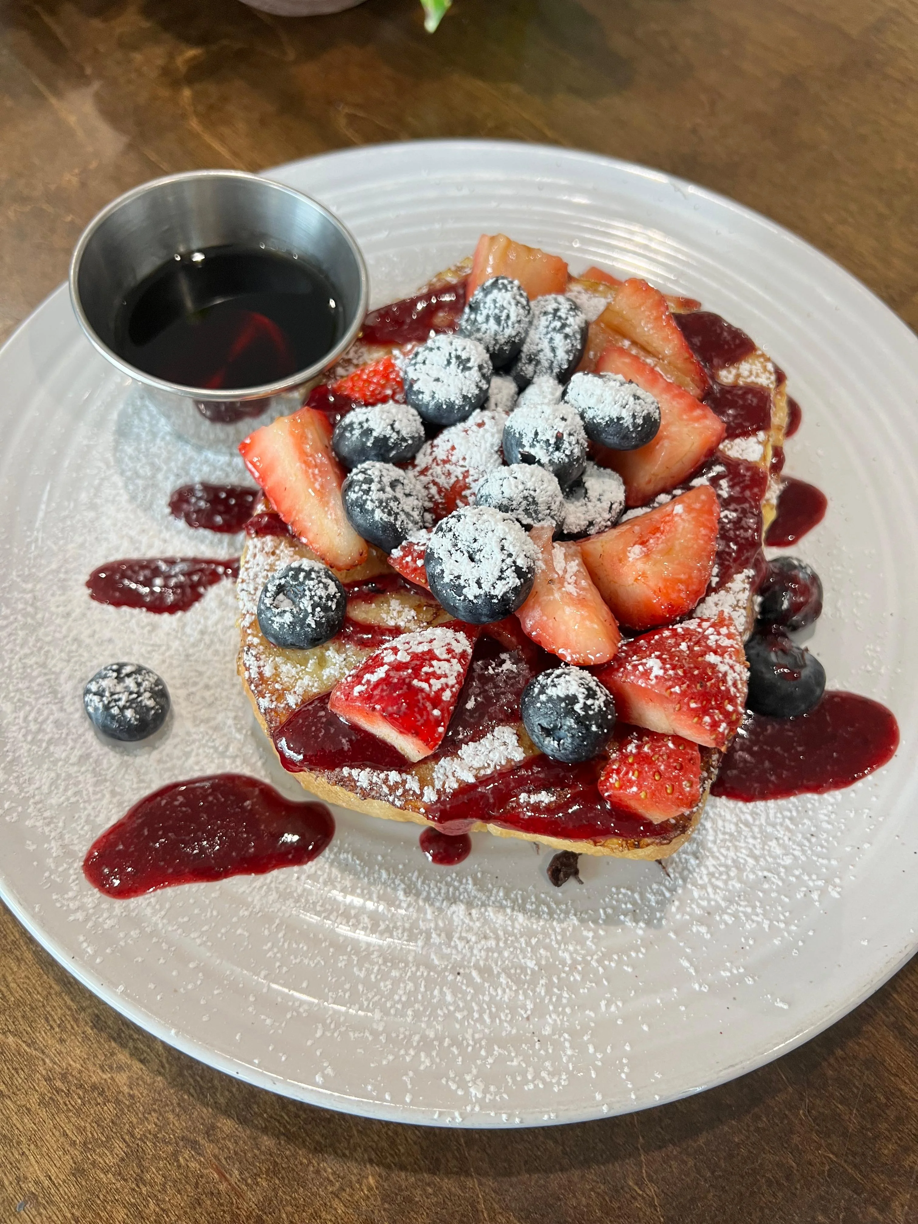 A delicious plate featuring a thick slice of French toast topped with fresh strawberries, blueberries, powdered sugar, and a drizzle of berry sauce, accompanied by a small cup of syrup.