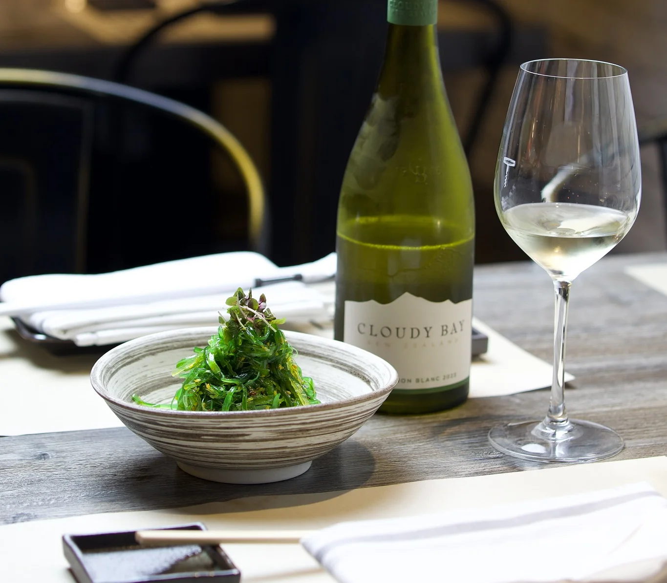 A bowl of vibrant green seaweed salad served on a wooden table alongside a bottle of Cloudy Bay white wine and a filled wine glass.