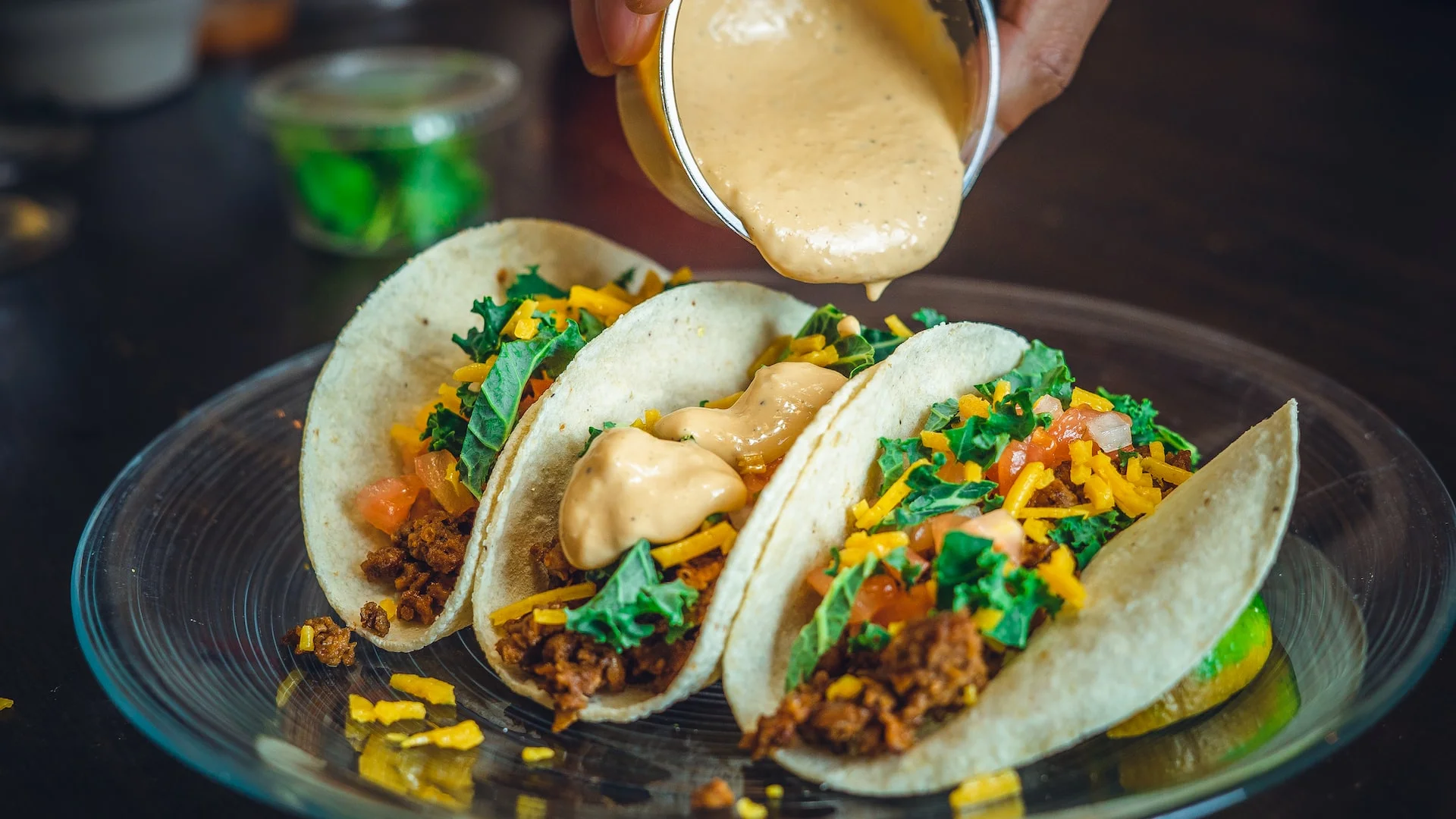 A plate of tacos filled with meat, vegetables, and topped with a creamy sauce. Fresh ingredients are visible alongside.