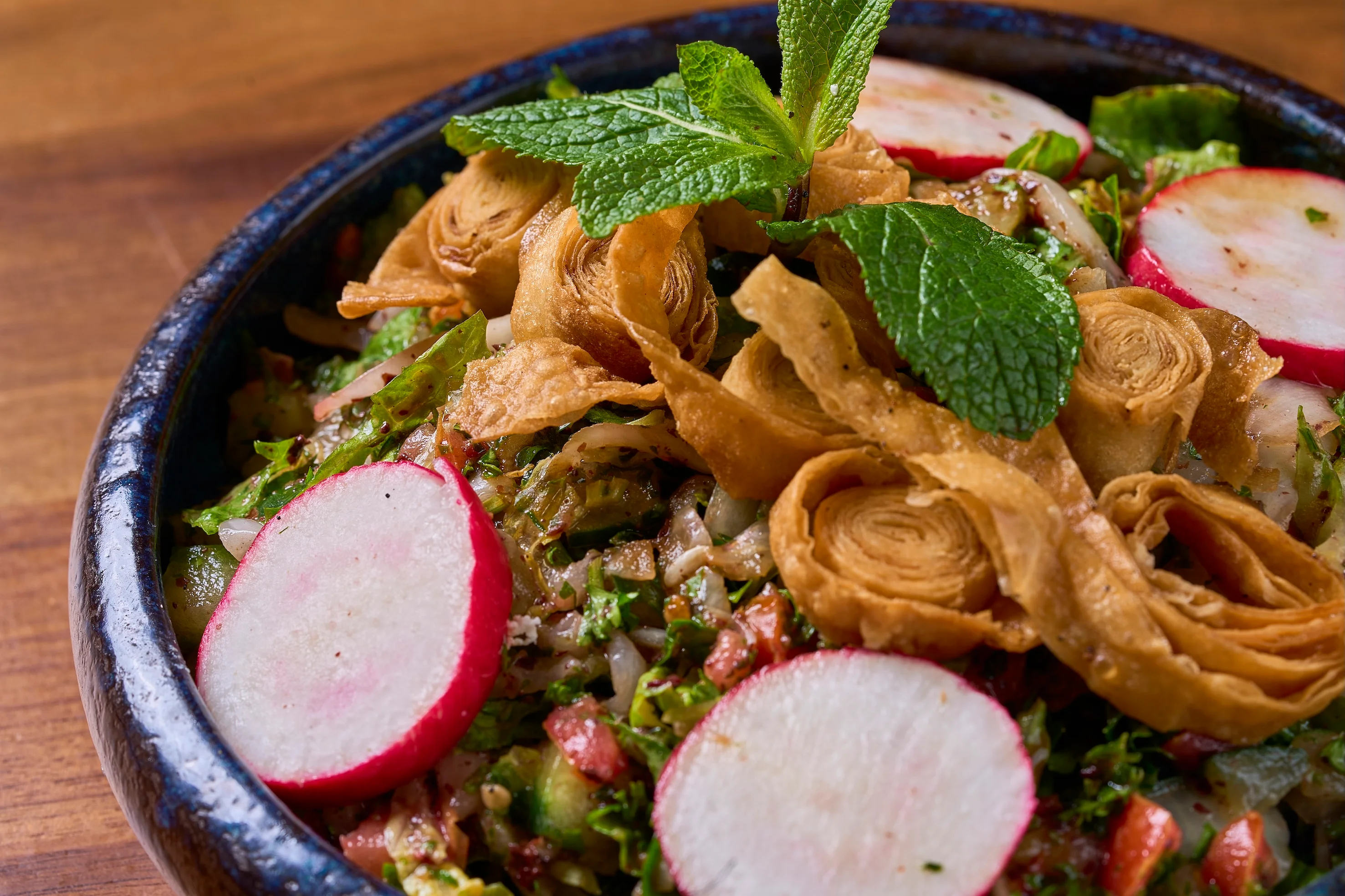 A vibrant salad in a black bowl featuring crisp, sliced radishes, fresh mint leaves, and golden-brown crispy rolls on a bed of mixed greens.