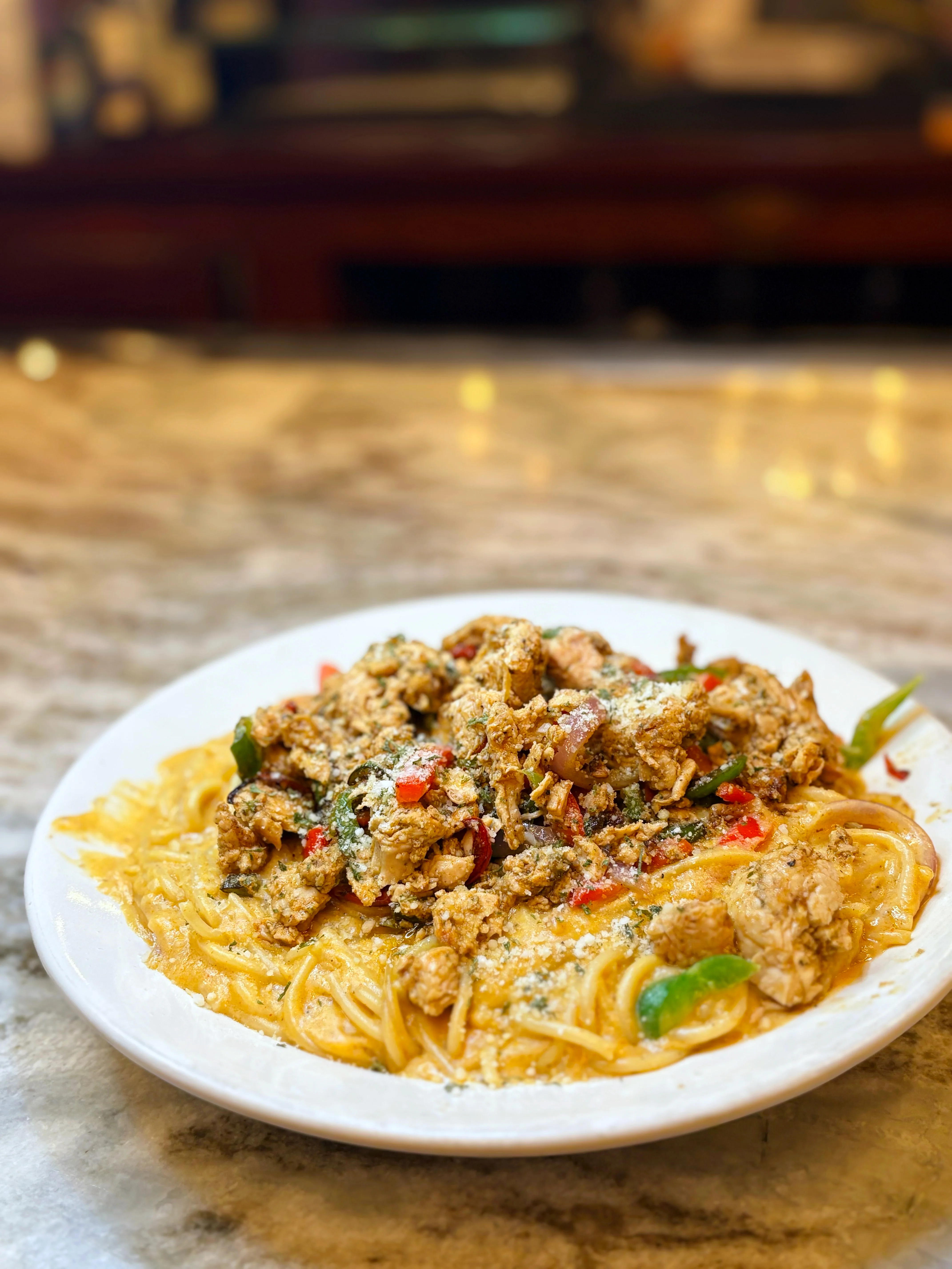 A plate of pasta topped with seasoned chicken and colorful vegetables, served with a sprinkle of cheese on a marble surface.