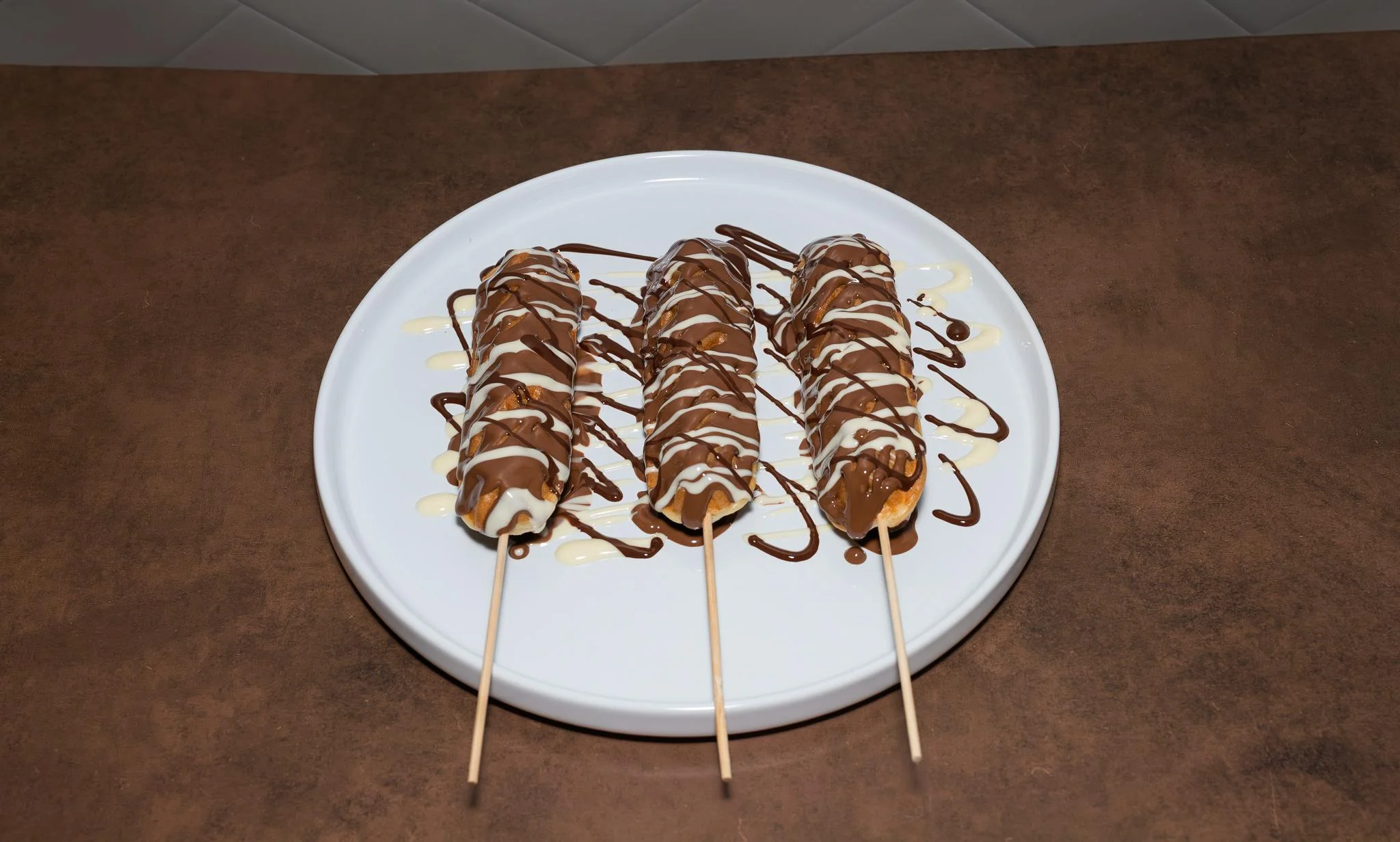Three dessert skewers featuring sweet treats, drizzled with chocolate and served on a white plate.