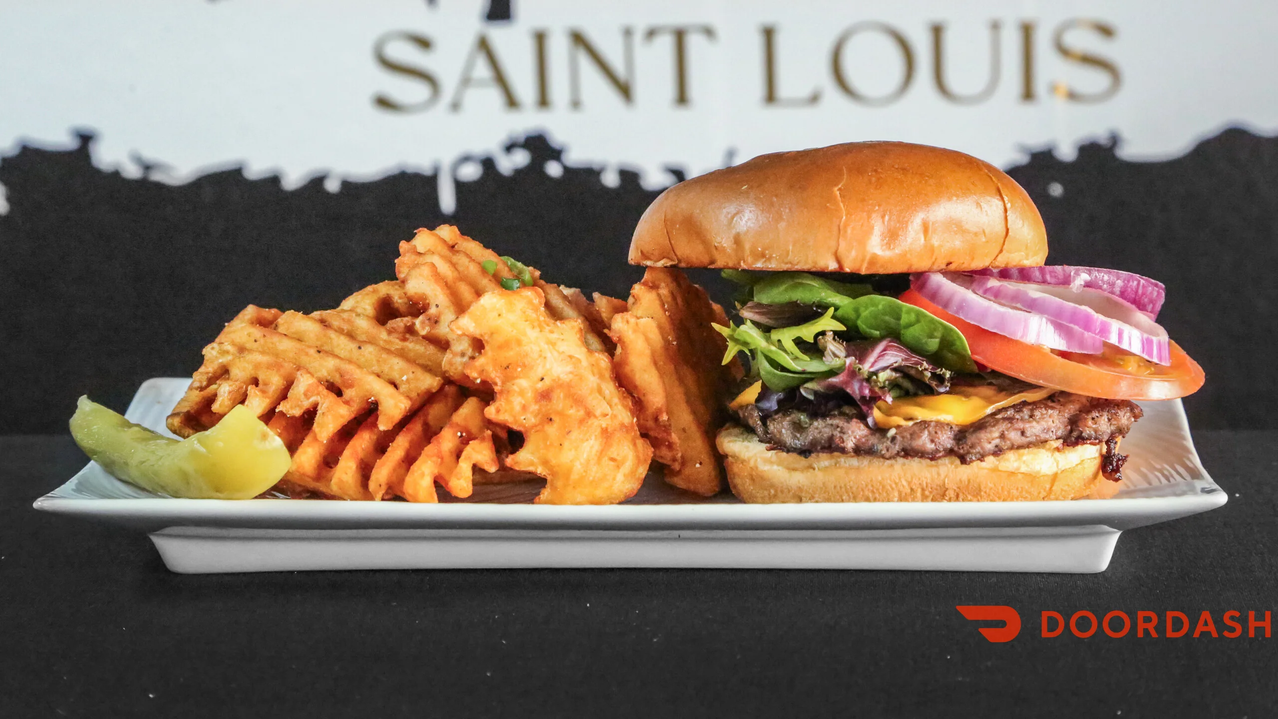 A plate featuring a hamburger with lettuce, tomato, and onion served alongside waffle fries and a slice of pickle.