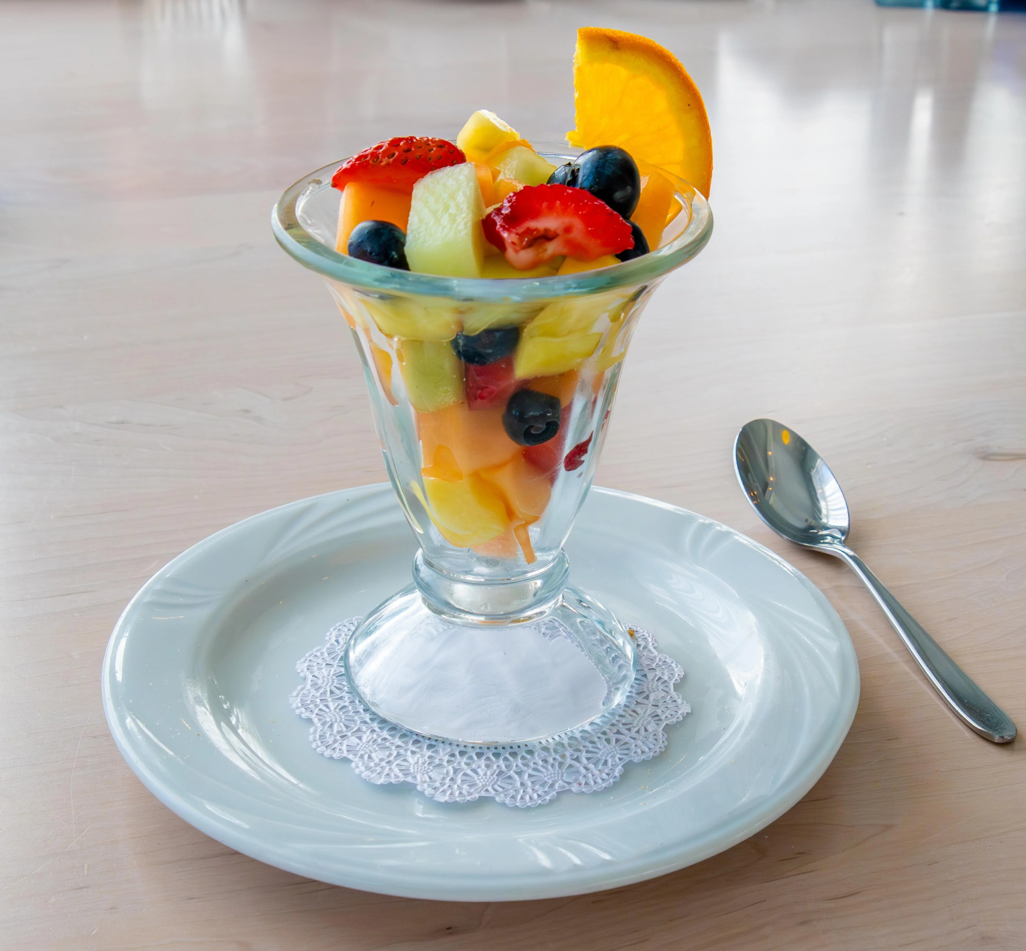 A glass dessert dish filled with a colorful assortment of fresh fruit, including melon, strawberries, and blueberries, garnished with a slice of orange.