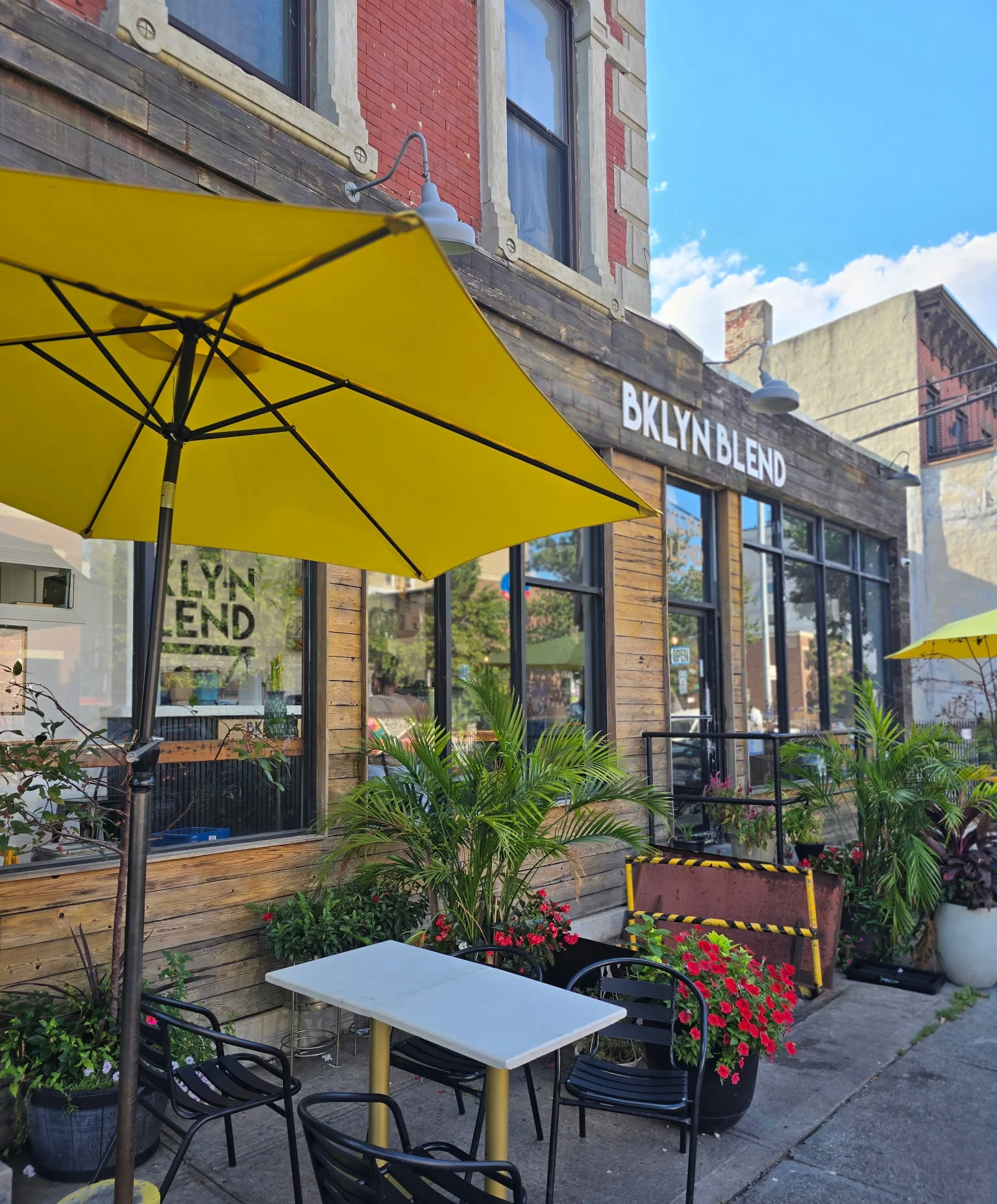Cozy cafe with rustic wooden exterior, large windows, and vibrant yellow umbrellas. "BKLYN BLEND" sign above. Lush greenery adds a fresh, inviting vibe.