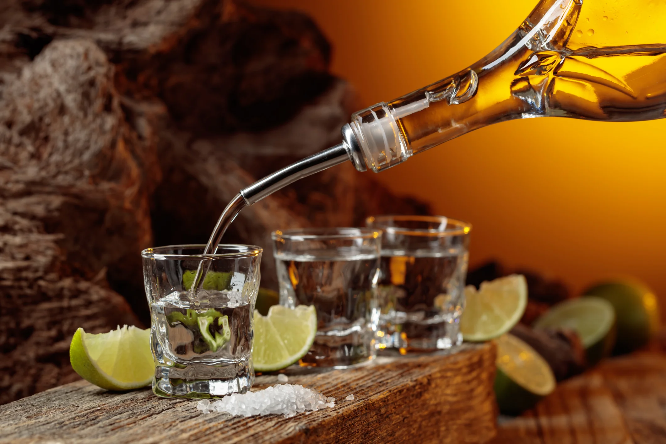 A bottle pouring liquor into shot glasses, accompanied by lime wedges and salt on a rustic wooden surface.