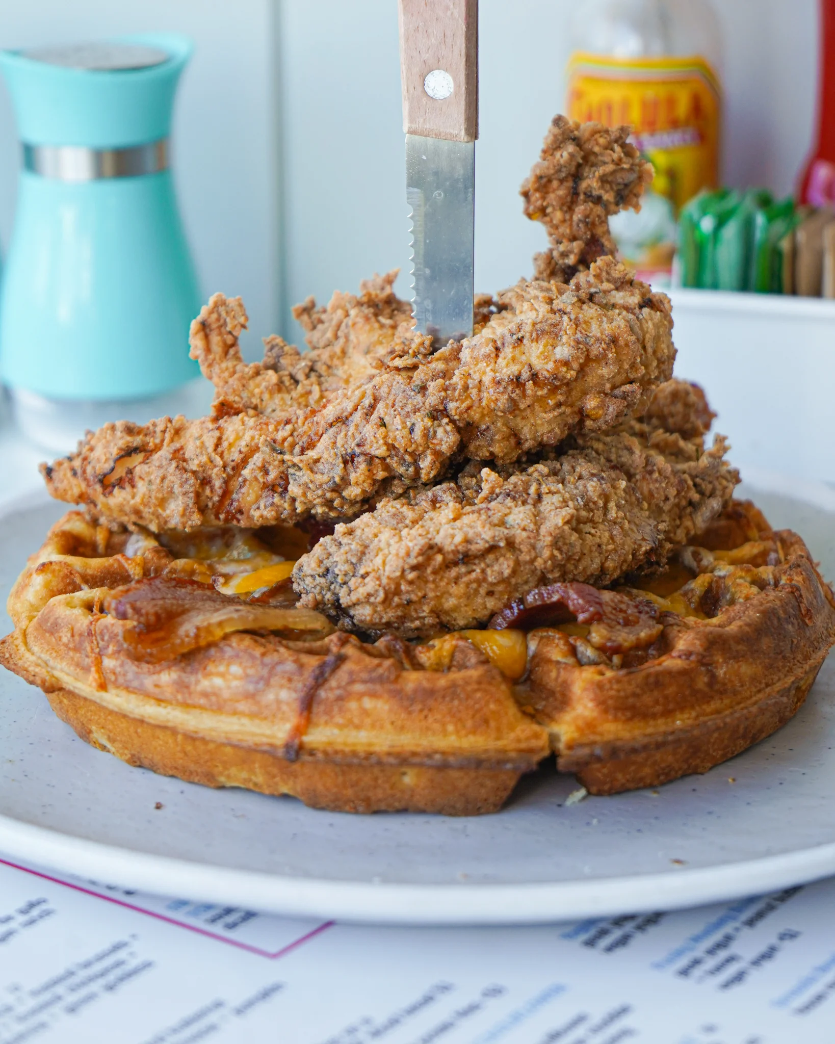 A hearty plate with a fluffy waffle topped with crispy fried chicken. The dish is served with a knife embedded in the chicken, showcasing the meal's generous portion.