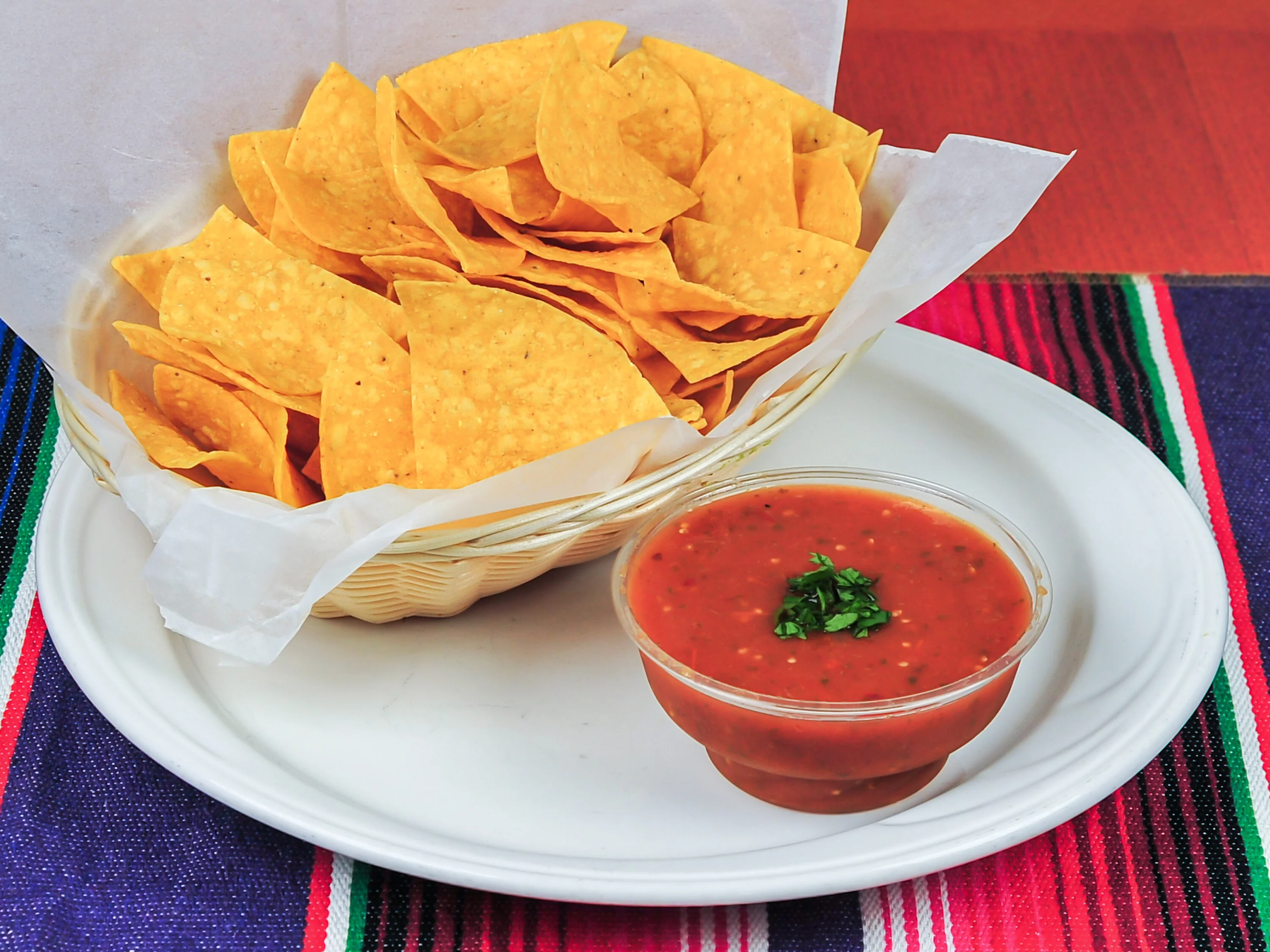A basket of golden tortilla chips accompanied by a small cup of red salsa, garnished with chopped cilantro, arranged on a decorative tablecloth.