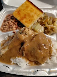 A takeout meal featuring rice with gravy, a piece of cornbread, black-eyed peas, and collard greens in a styrofoam container.