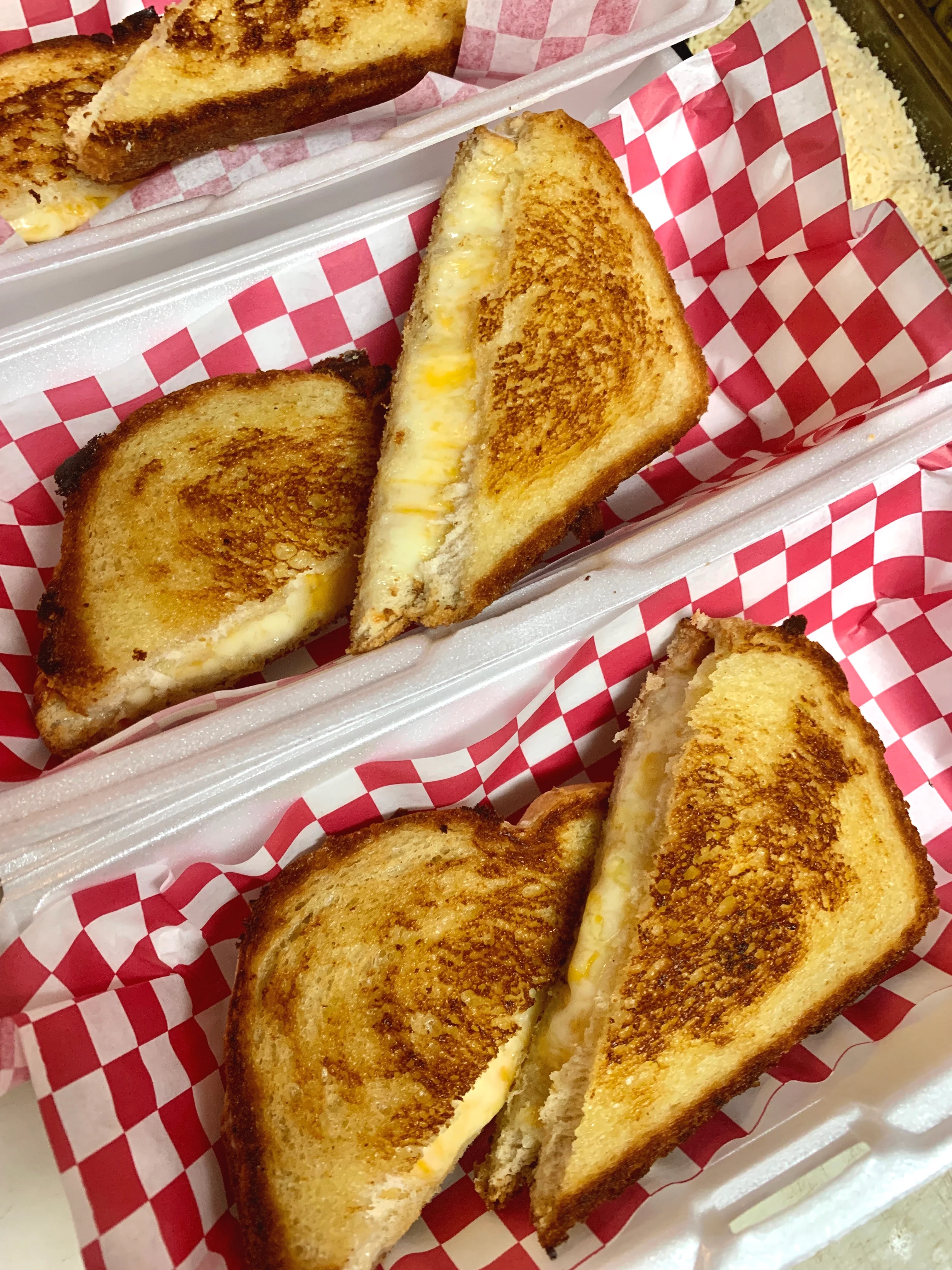 Golden grilled cheese sandwiches with melted cheddar and mozzarella, served in foam takeout containers lined with red and white checkered paper.