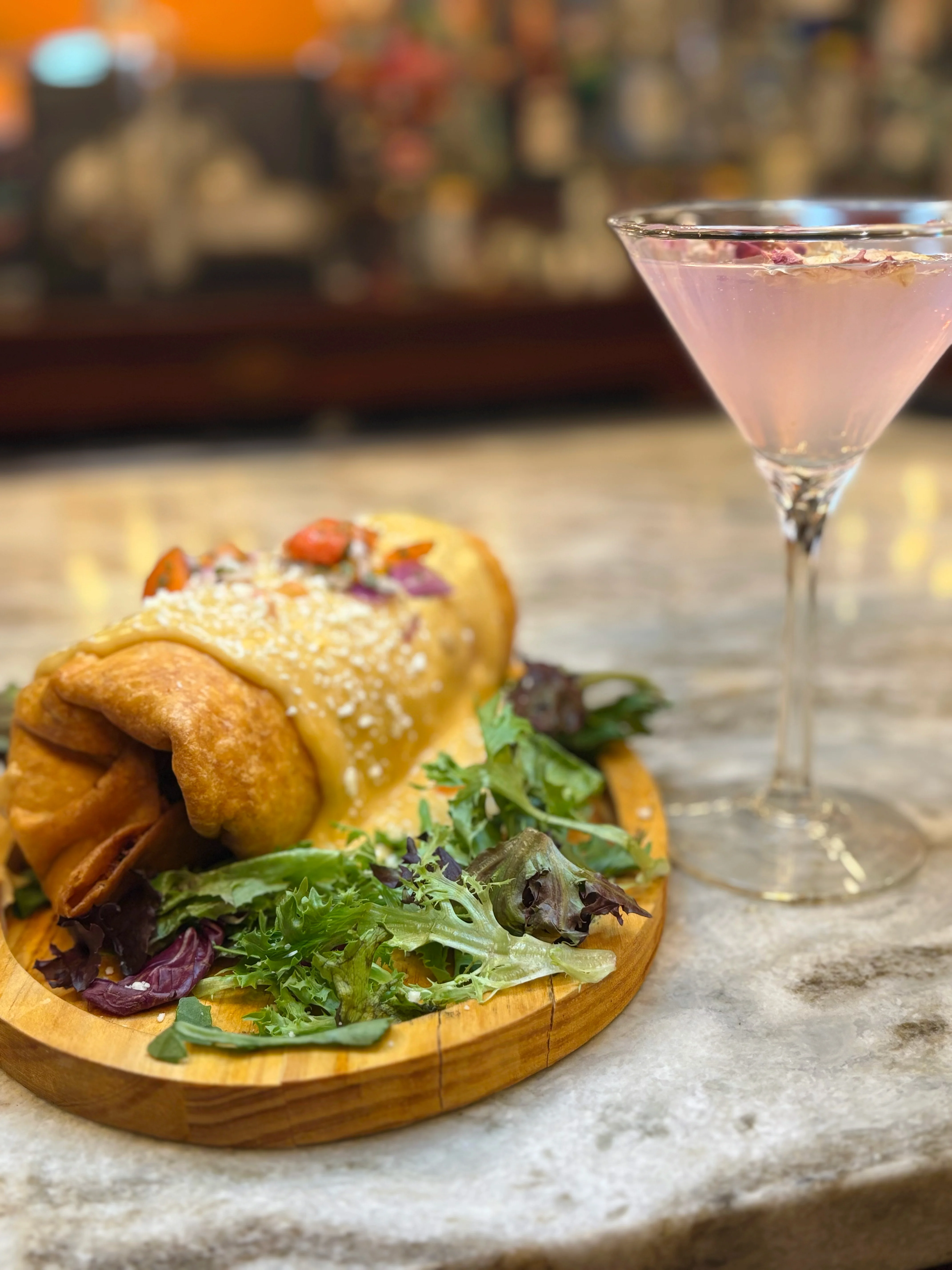 A large rolled tortilla filled with ingredients, garnished with sesame seeds, served alongside a pink cocktail in a martini glass.