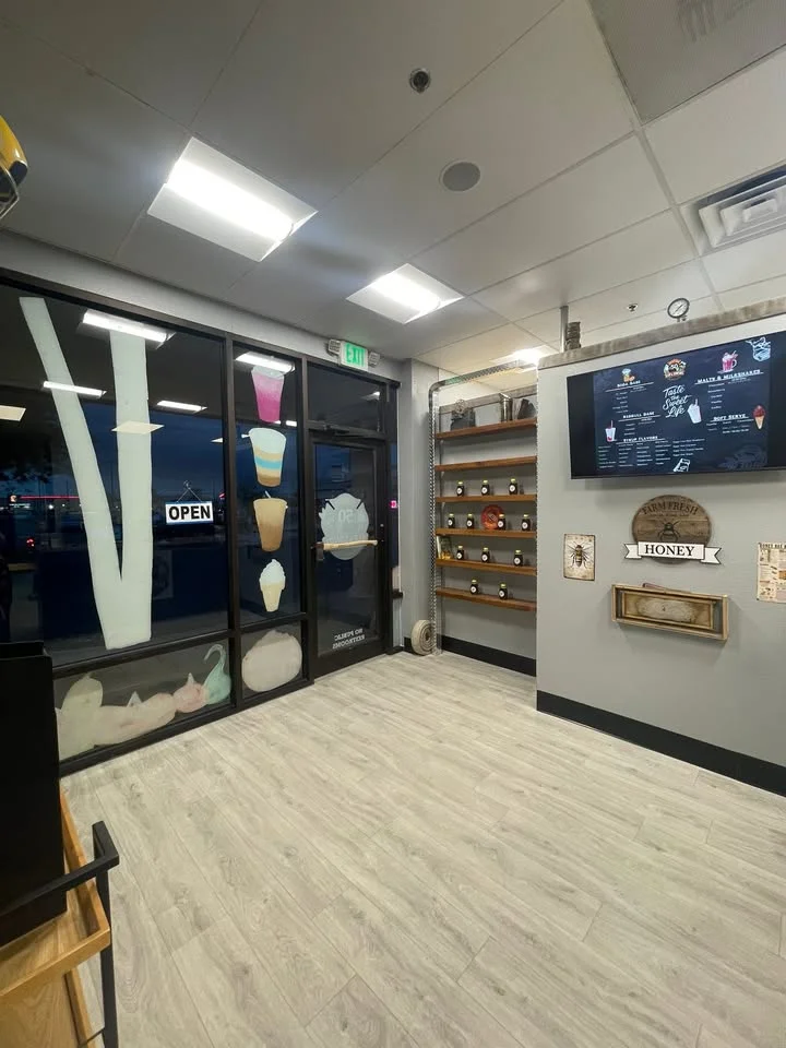 An interior view of a beverage shop with open signage, showcasing the menu on the wall and shelves for product display.