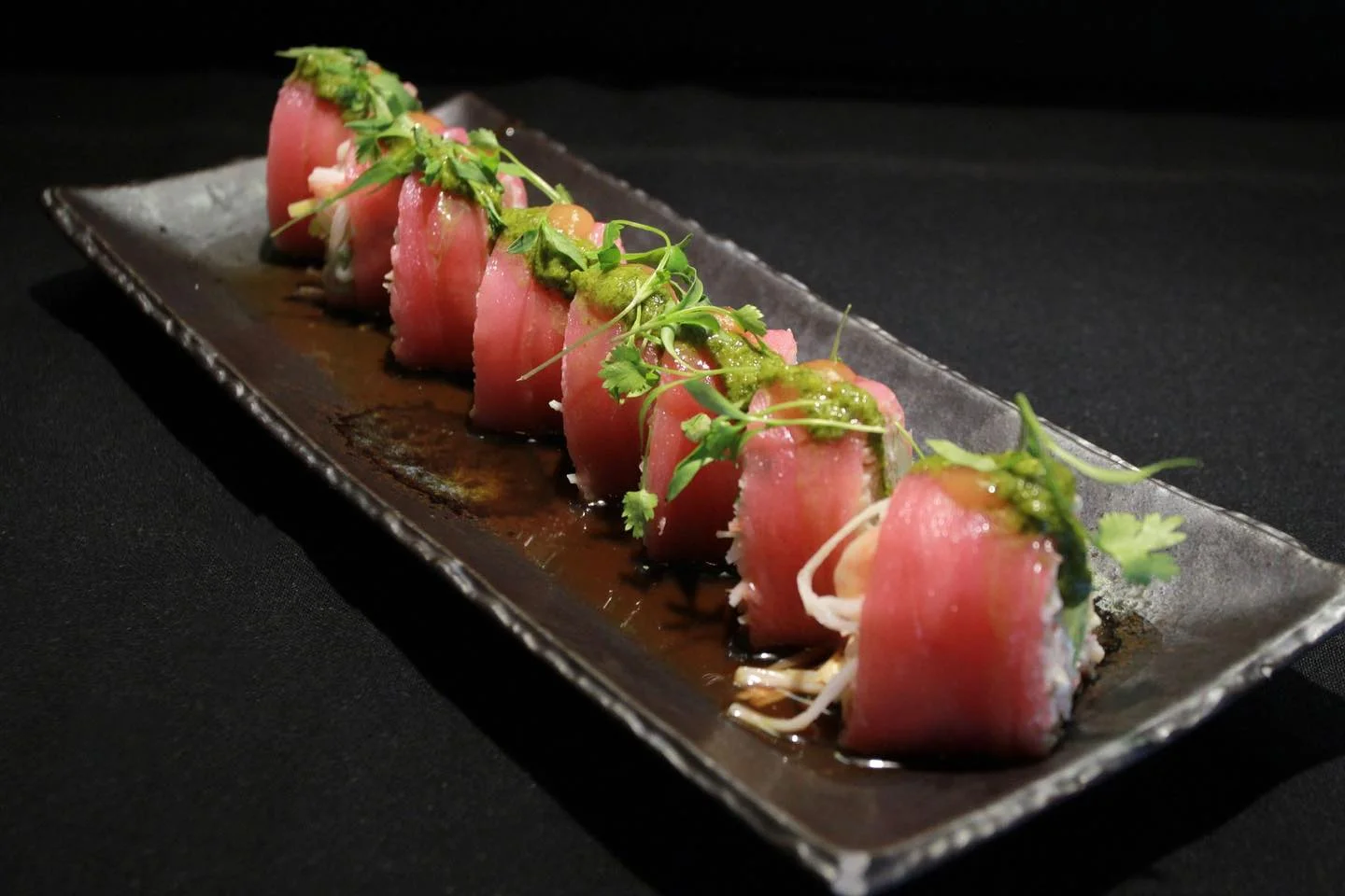 Sushi rolls wrapped in fresh tuna, topped with green herb sauce and microgreens, served on a long dark plate with light sauce.