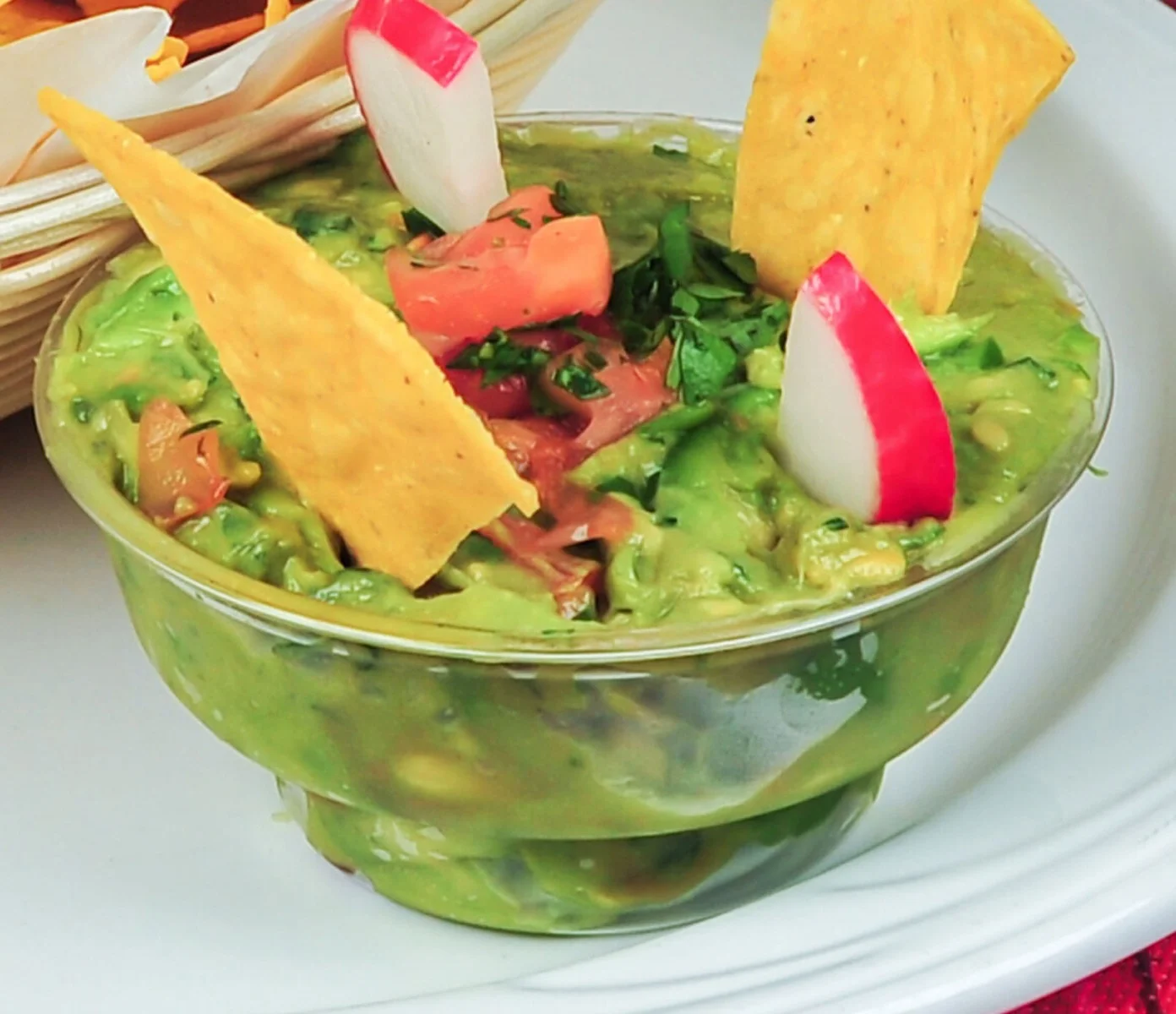 A bowl of guacamole garnished with two tortilla chips, diced tomatoes, cilantro, and slices of radish, served with a side of tortilla chips.