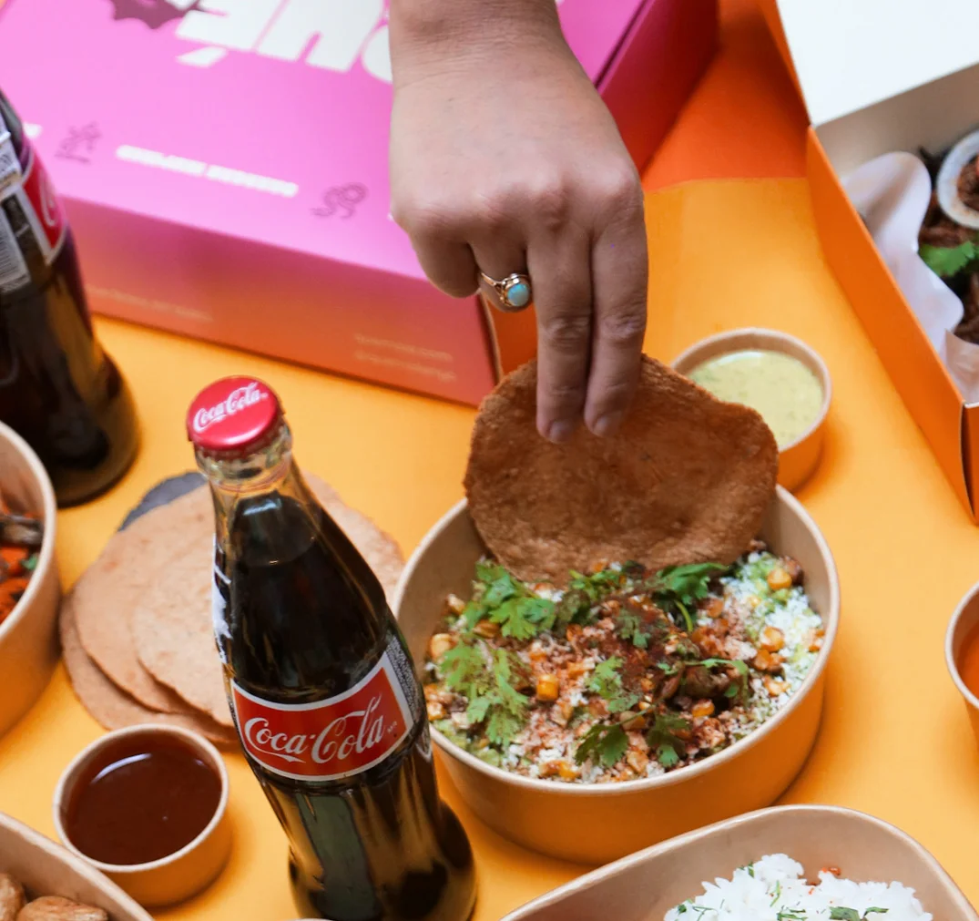 A hand dipping a crispy tortilla into a colorful bowl of Mexican food, surrounded by drinks and toppings.