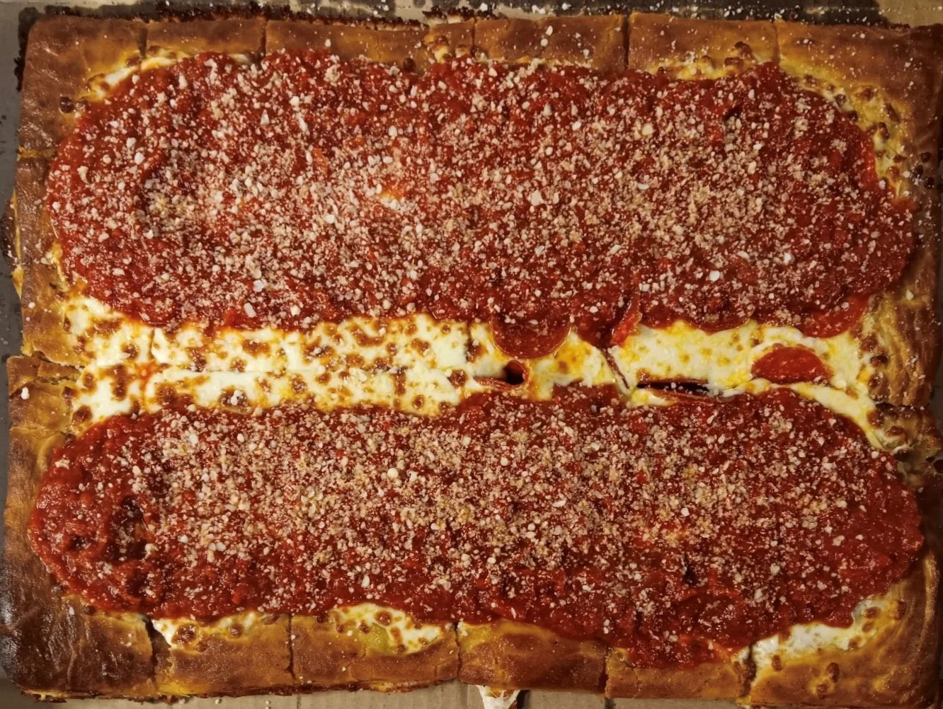 A rectangular pizza topped with sauce, cheese, and a sprinkle of herbs or breadcrumbs. The edges are golden brown.