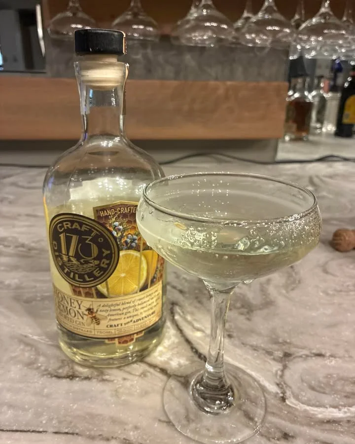 A glass of clear sparkling cocktail beside a bottle of handcrafted gin, set on a marble surface.