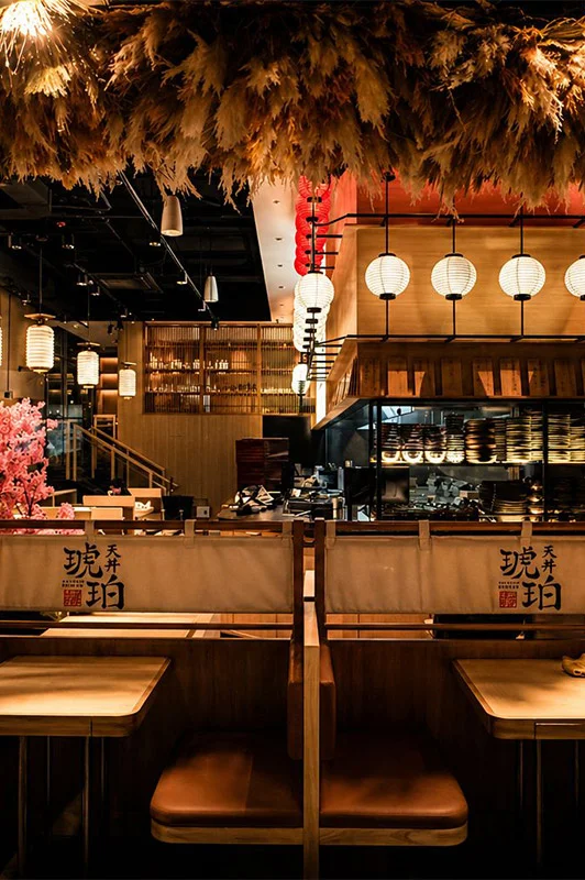 A warmly lit view of the Tendon Kohaku dining room showing two wooden booths with branded canvas dividers. Above the booth is a dense canopy of fluffy pampas grass creates a dramatic ceiling texture, accented by hanging white globe lanterns and a sleek open kitchen in the background.