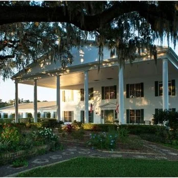 A large white house with a porch surrounded by greenery and flower beds.