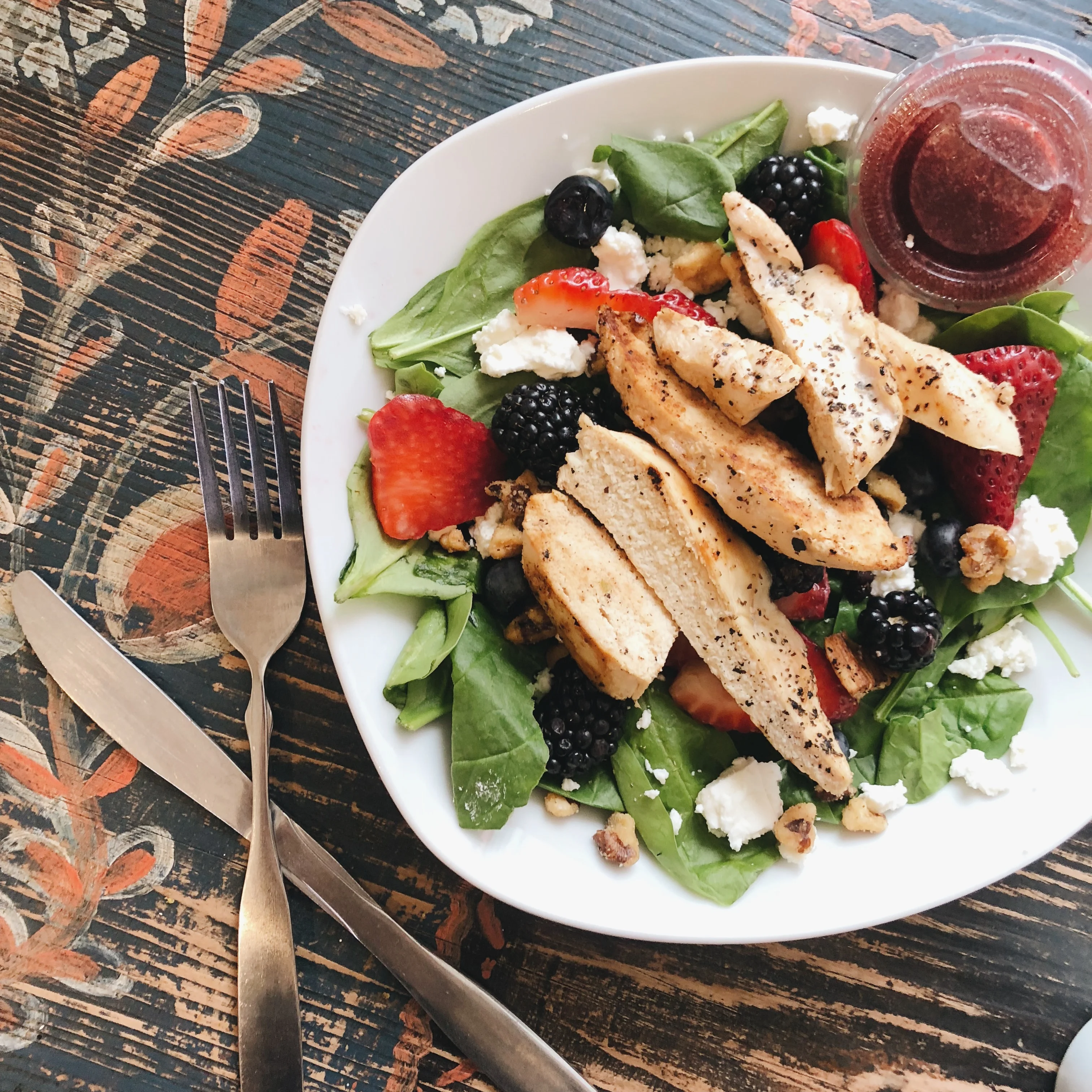 A colorful salad featuring grilled chicken, fresh spinach, assorted berries, and crumbled cheese, served with a side of dressing.