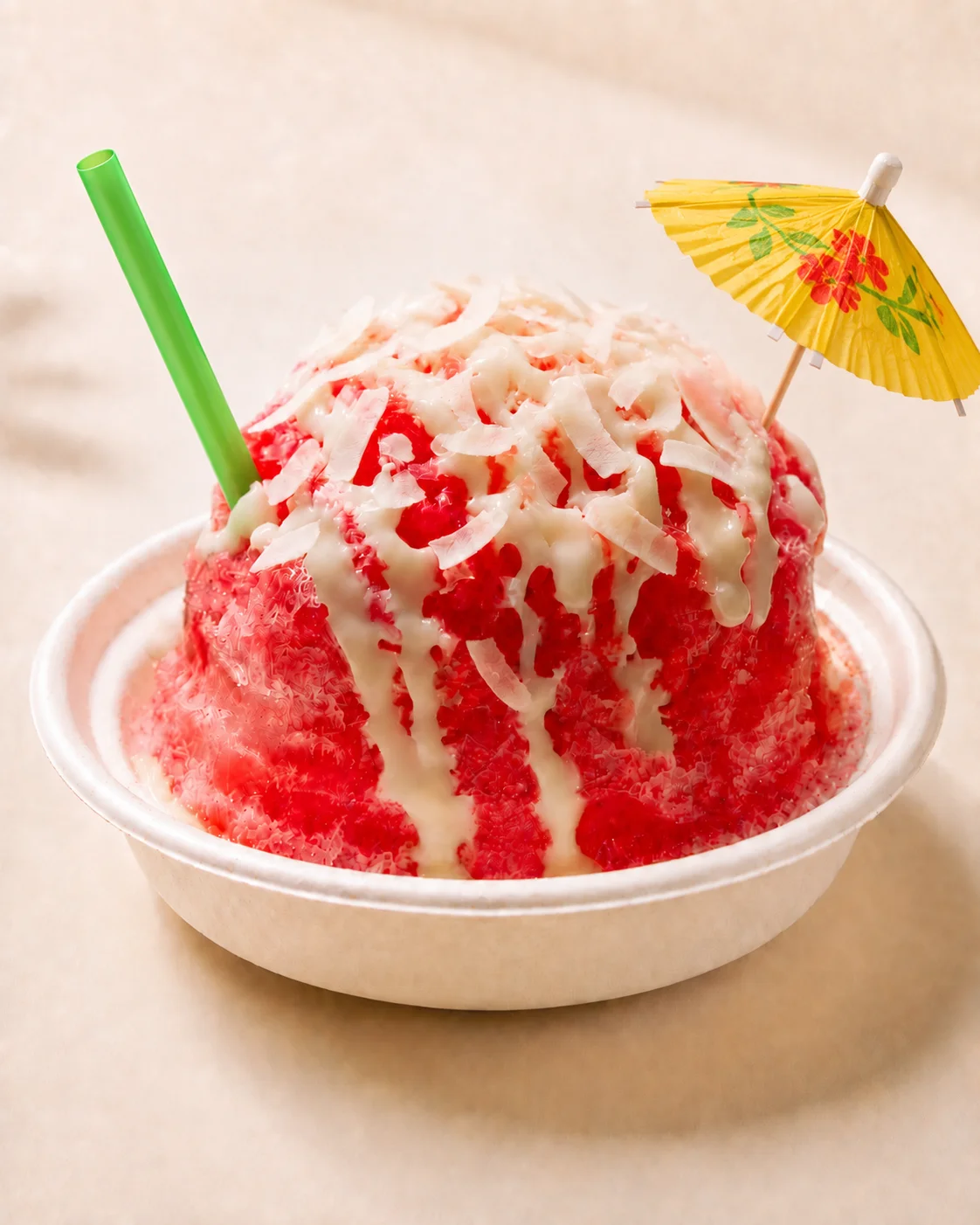 A bowl of colorful shaved ice with a fruity flavor, topped with coconut shavings and accompanied by a small decorative umbrella and straw.