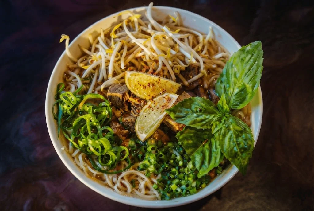 A bowl of beef noodle soup with fresh herbs and vegetables.