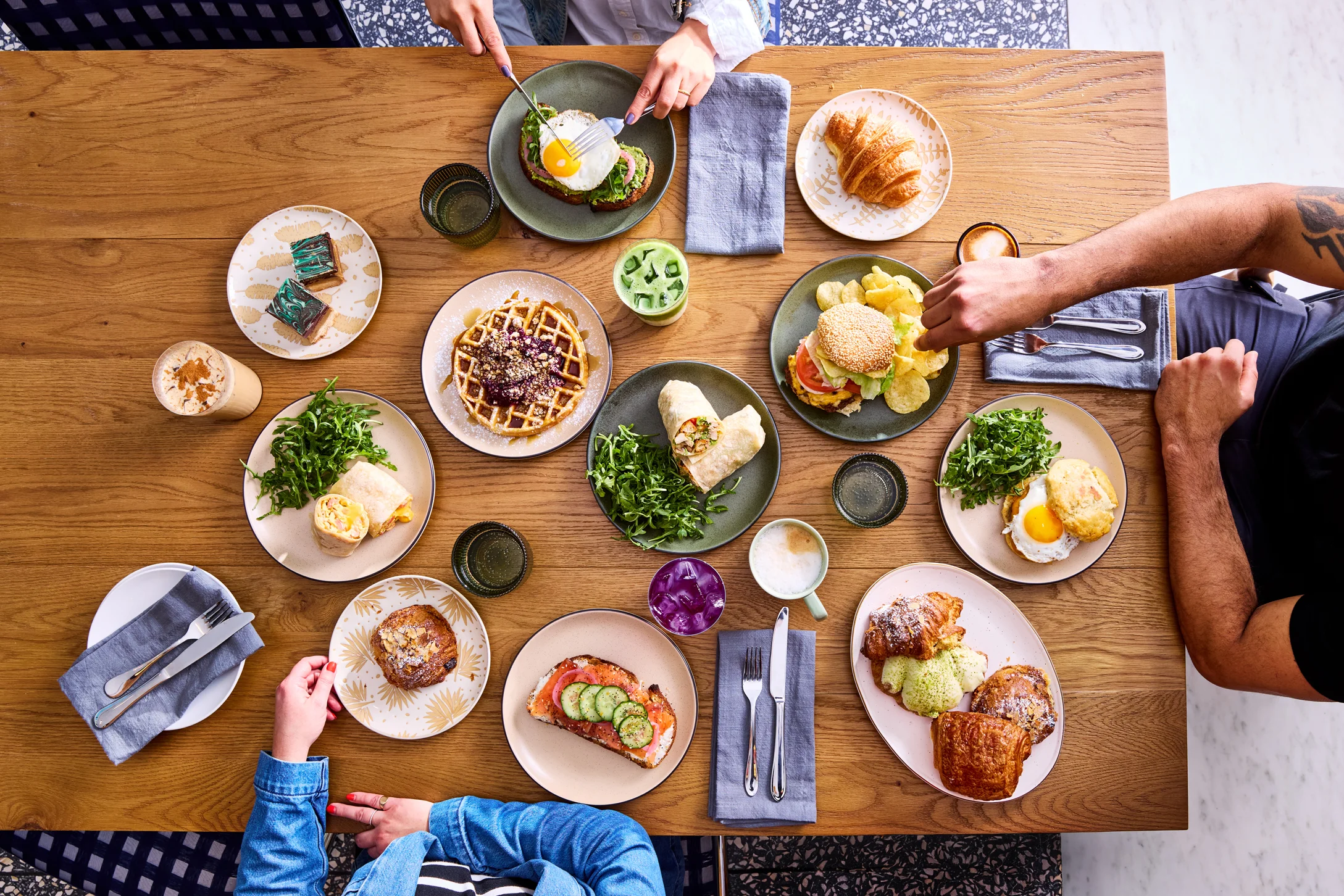 An overhead view of a diverse breakfast spread including eggs, waffles, sandwiches, salads, and drinks arranged on a wooden table.