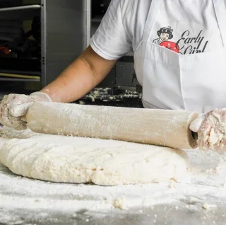 A person preparing dough, holding a piece while shaping it. The work surface is covered in flour, and a rolling pin is nearby.
