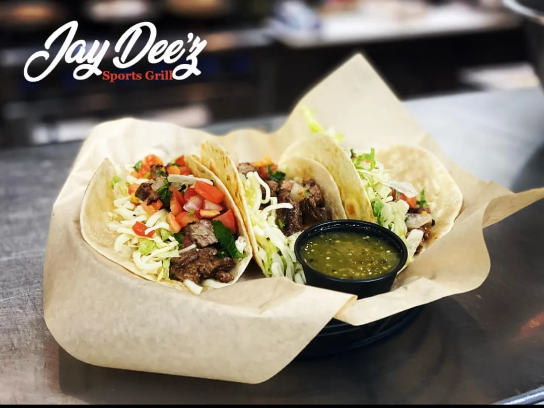 Three soft tacos filled with meat and topped with lettuce, tomatoes, and cheese, accompanied by a small cup of green salsa.
