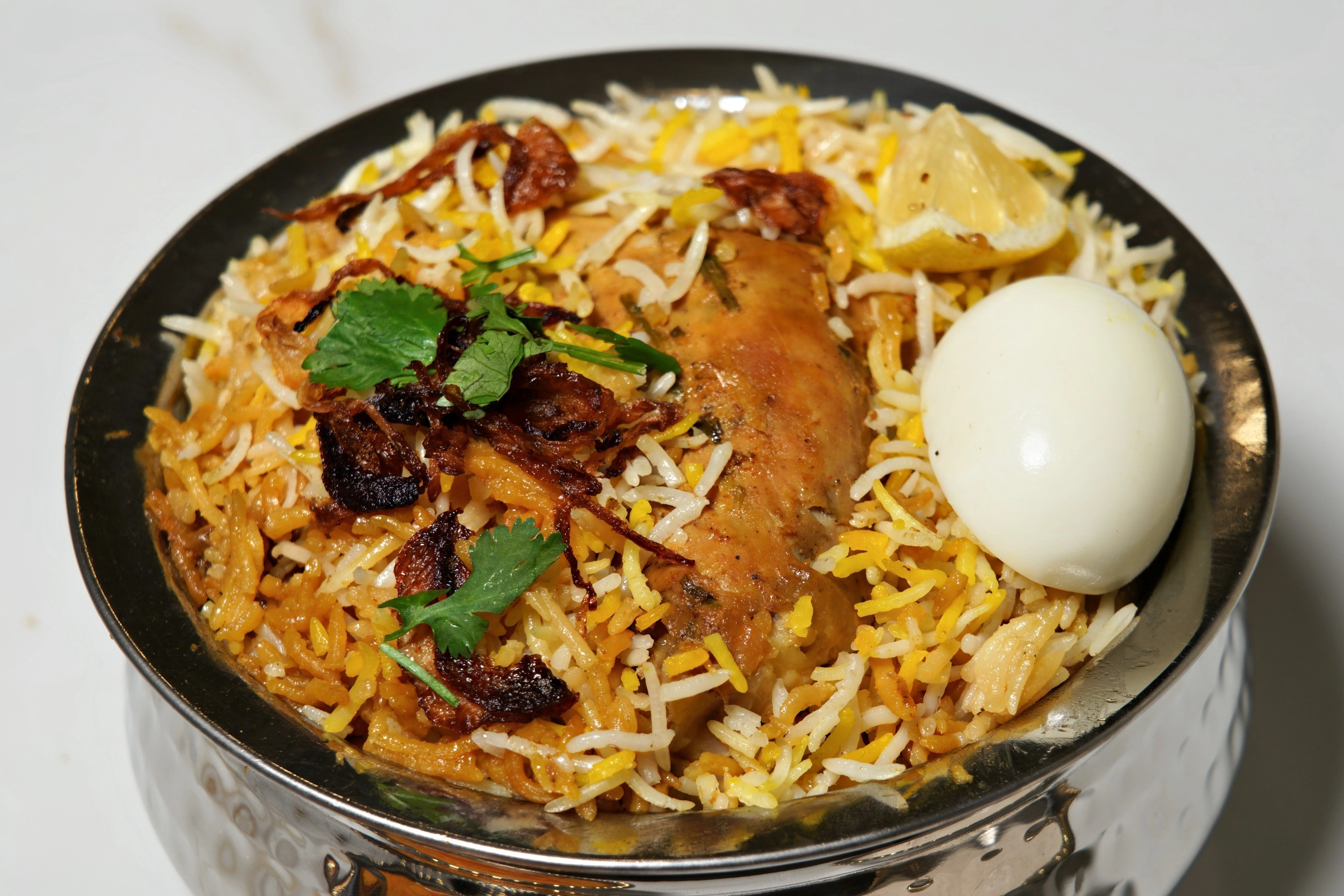 A bowl of biryani featuring layers of aromatic rice, tender chicken, a boiled egg, and garnished with fried onions and fresh herbs.
