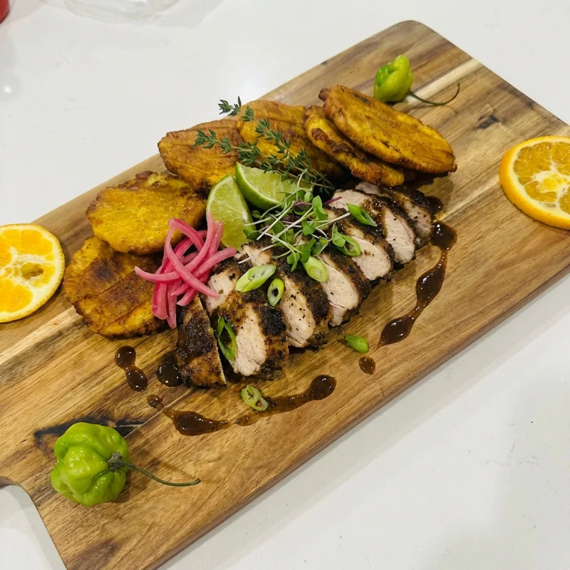 A wooden platter presenting sliced grilled chicken, fried plantains, lime, and fresh herbs, garnished with colorful toppings.