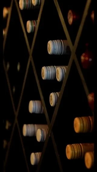 A collection of wine bottles stored horizontally in a dark, diamond-patterned wooden wine rack, highlighting the various colored caps and seals.