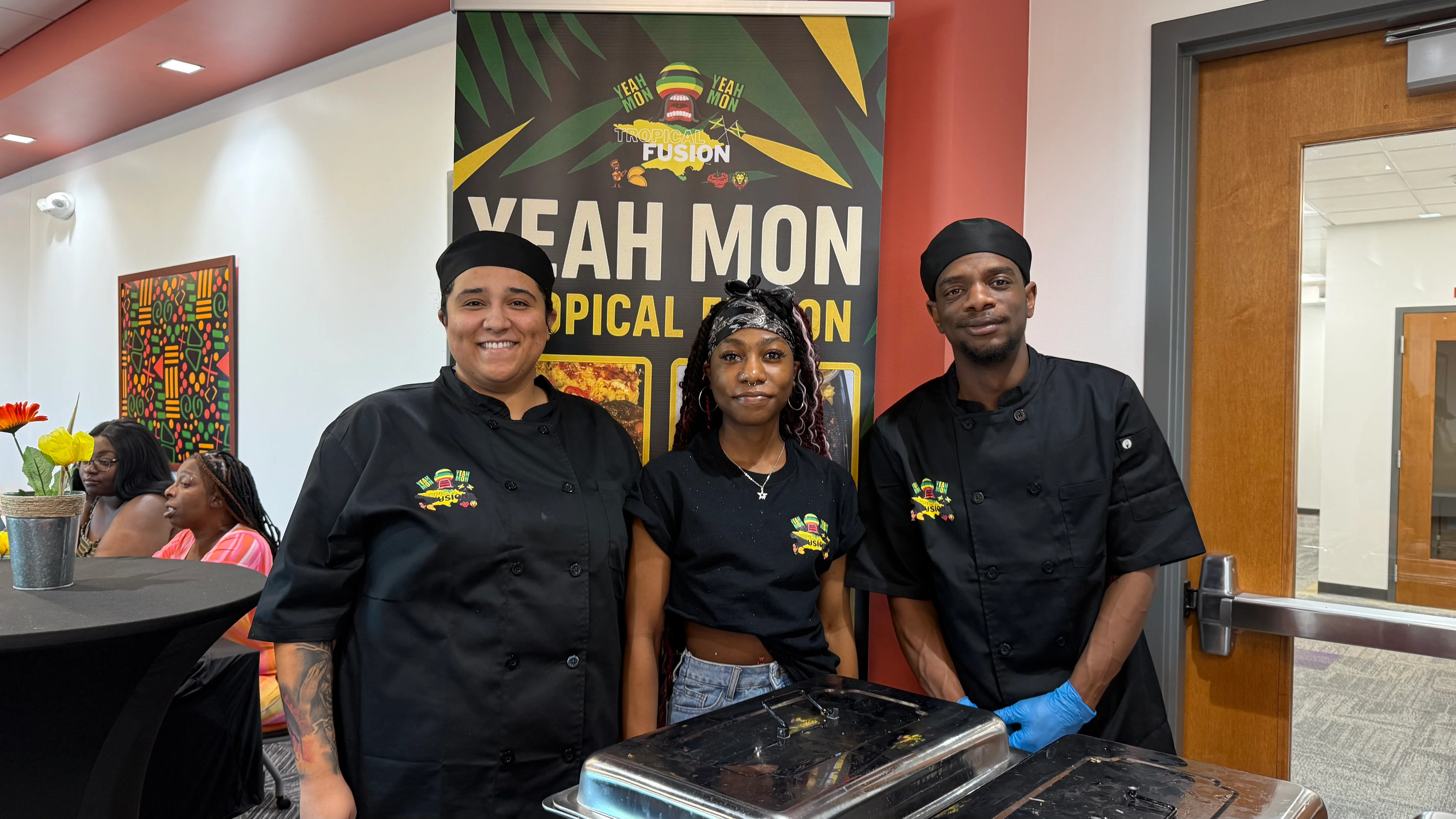 A group of chefs posing in front of a colorful promotional banner. They are dressed in black chef uniforms and are smiling at the camera, indicating a lively atmosphere.