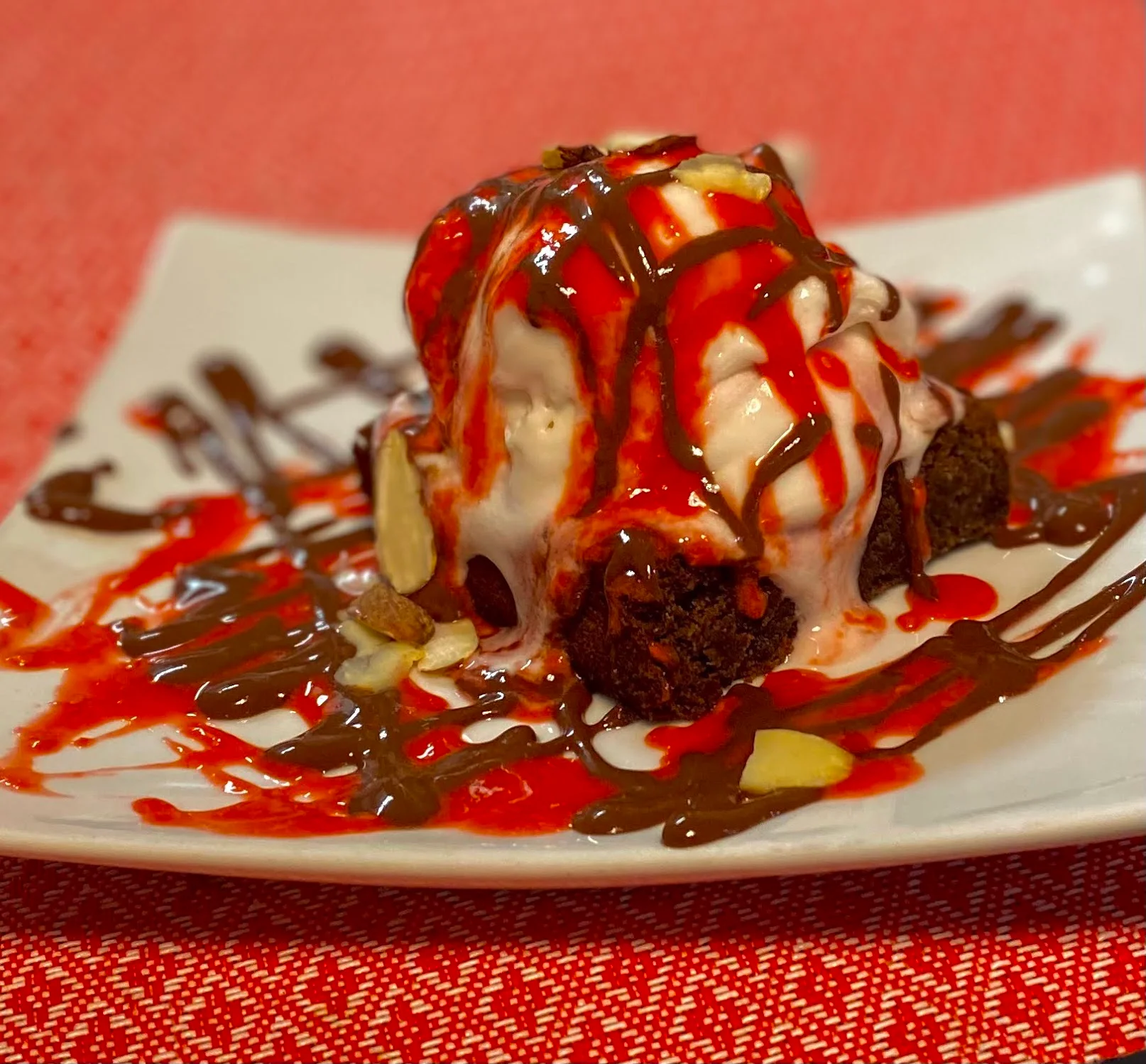 A dessert plate featuring a warm brownie topped with ice cream, drizzled with chocolate and strawberry sauce, and garnished with almonds.