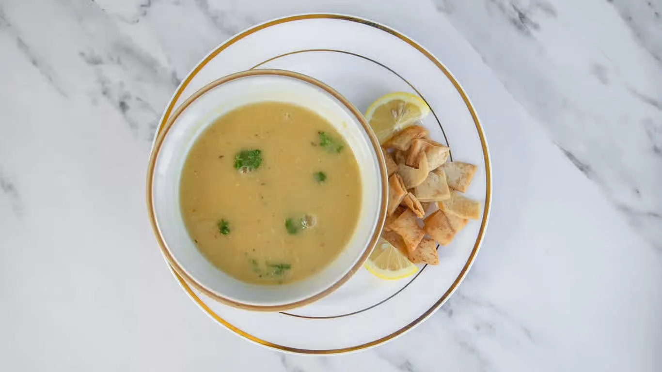 A warm bowl of soup with visible vegetables, served with golden crispy pita bread and a slice of lemon.