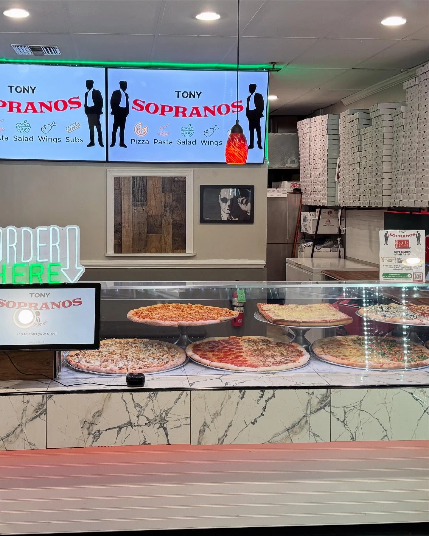 A pizza shop counter with various pizzas displayed under glass. The shop features signage that highlights its menu items.