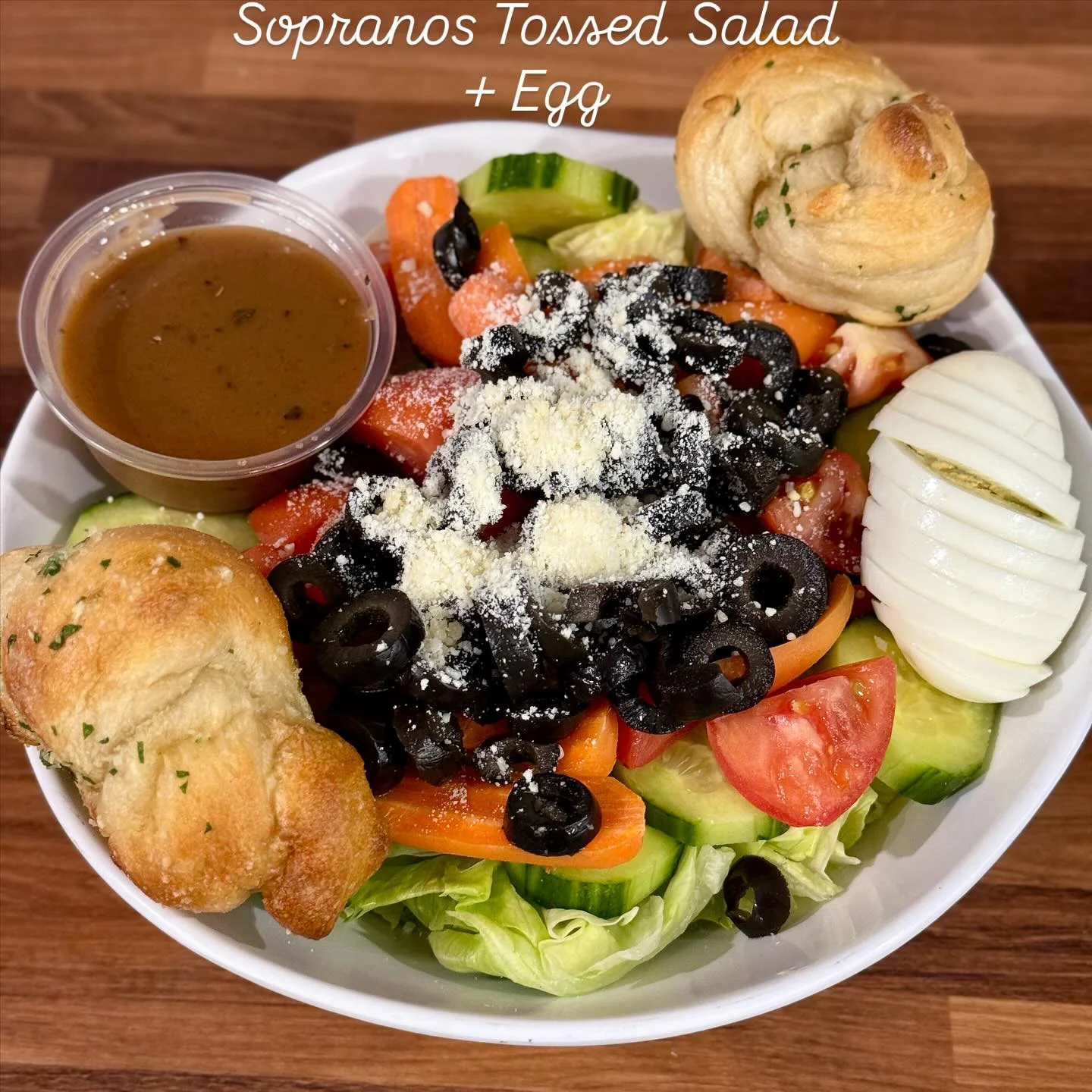 A colorful salad with fresh vegetables, olives, sliced egg, and a side of dressing, served with a bread roll.