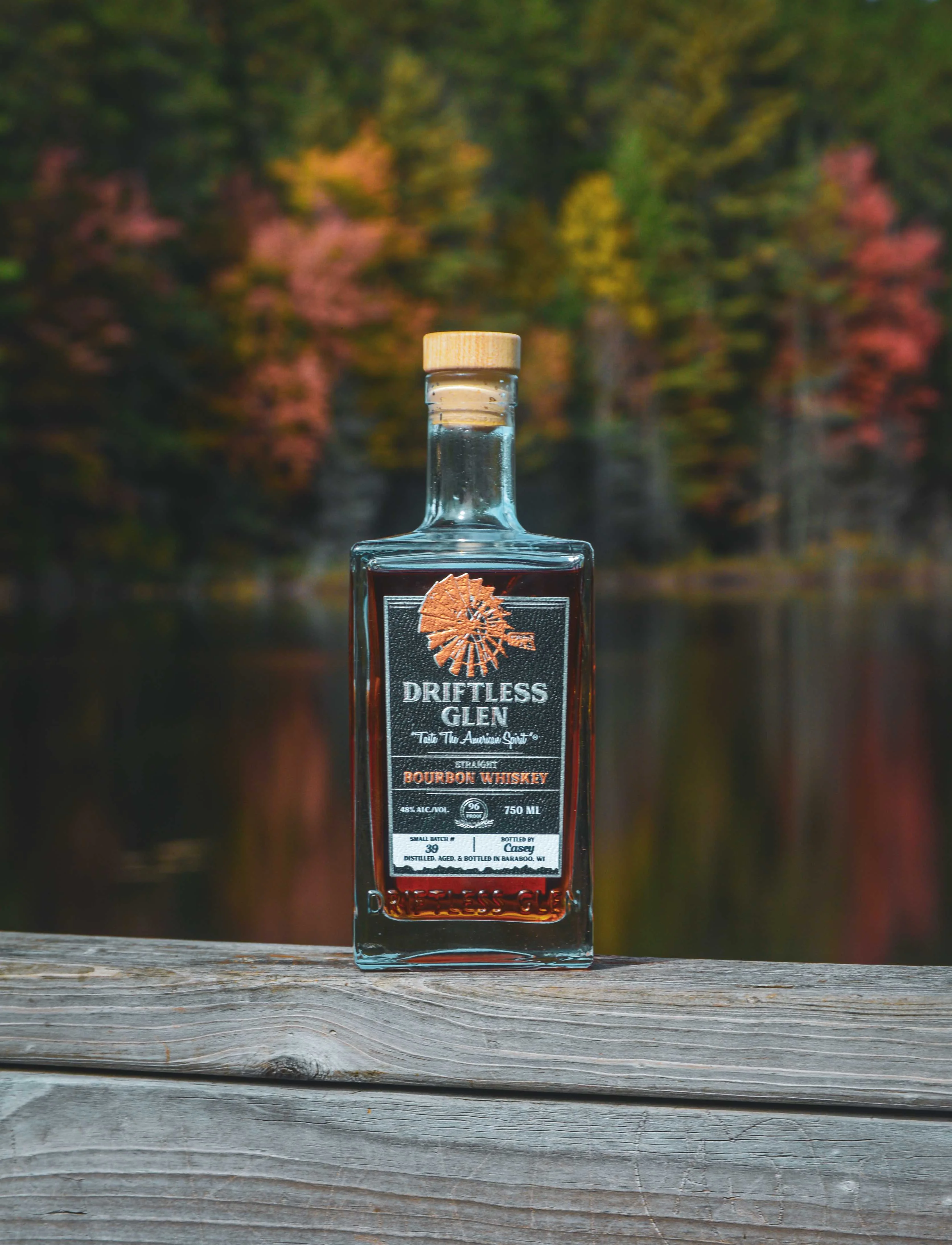 A bottle of Driftless Glen's small batch bourbon sits in front of a lake with fall colors on the leaves behind it and reflected into the water 