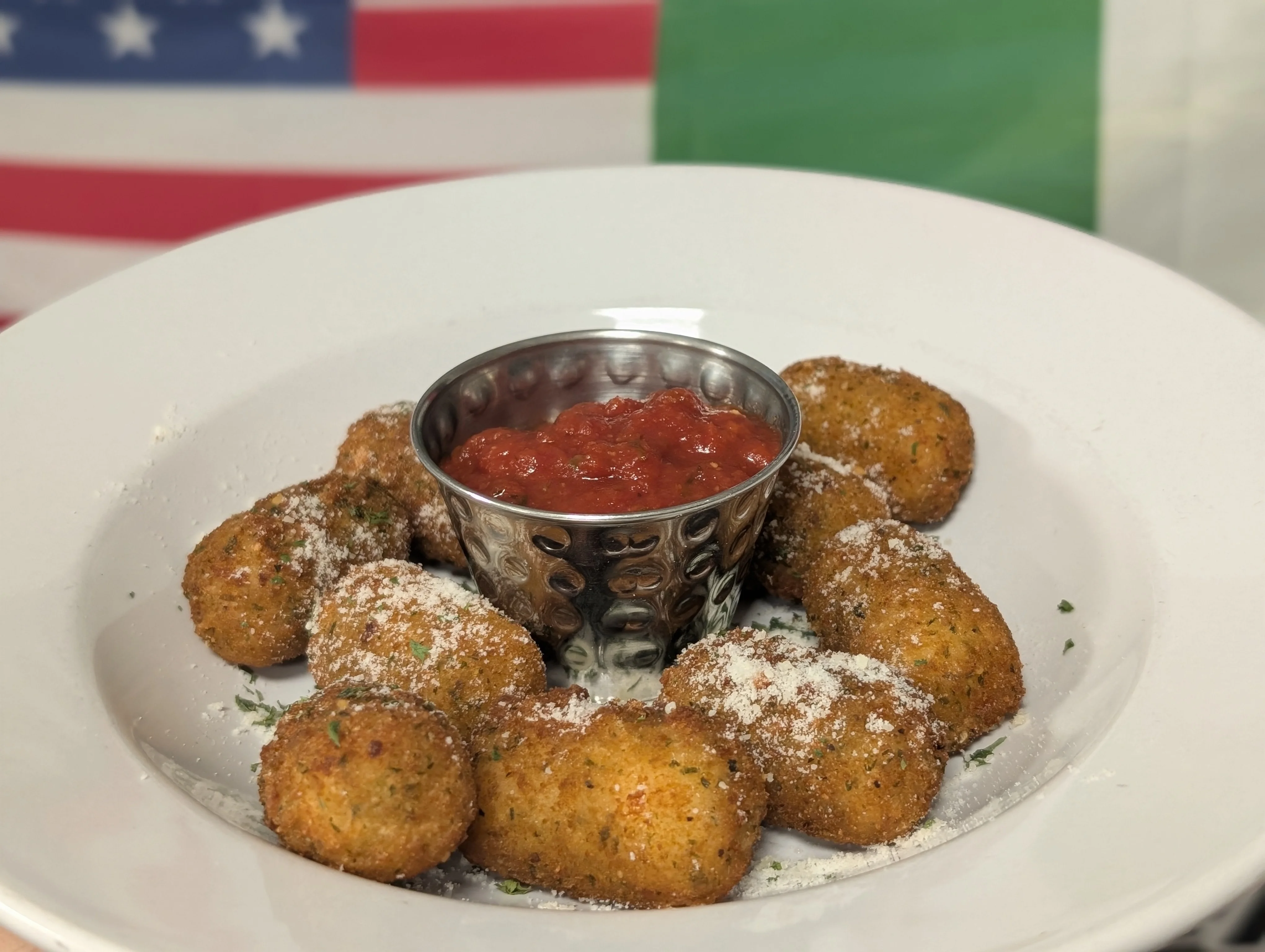Fried cheese balls sprinkled with parmesan and served with marinara sauce in a silver cup, with a background featuring the American and Italian flags.