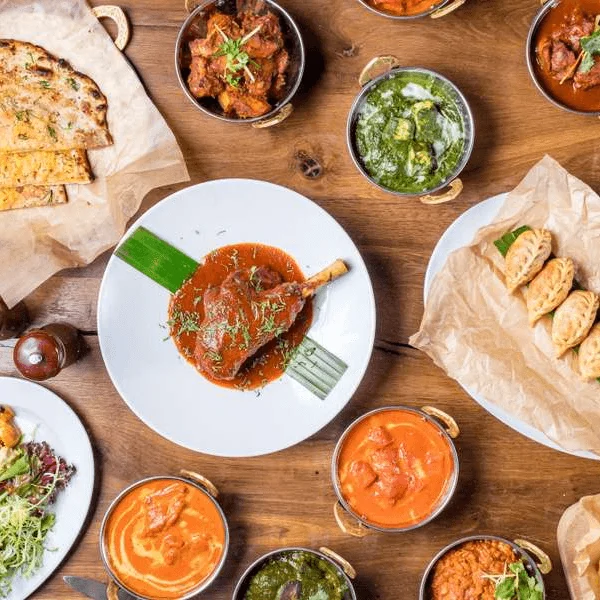 Spread of various Indian curries, naan, and a lamb shank on a table.