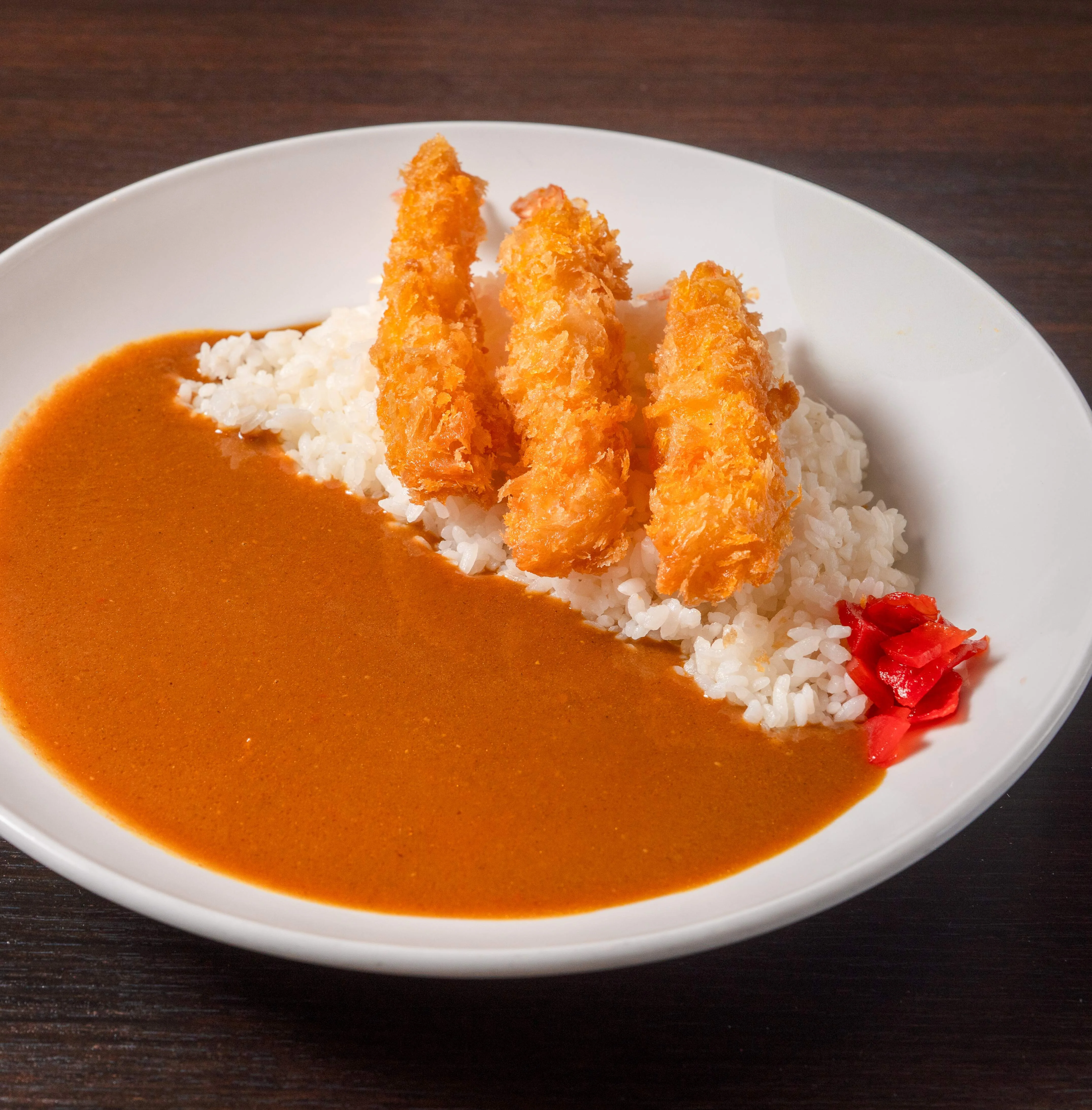 A plate featuring rice topped with curry and crispy shrimp, garnished with pickled vegetables.