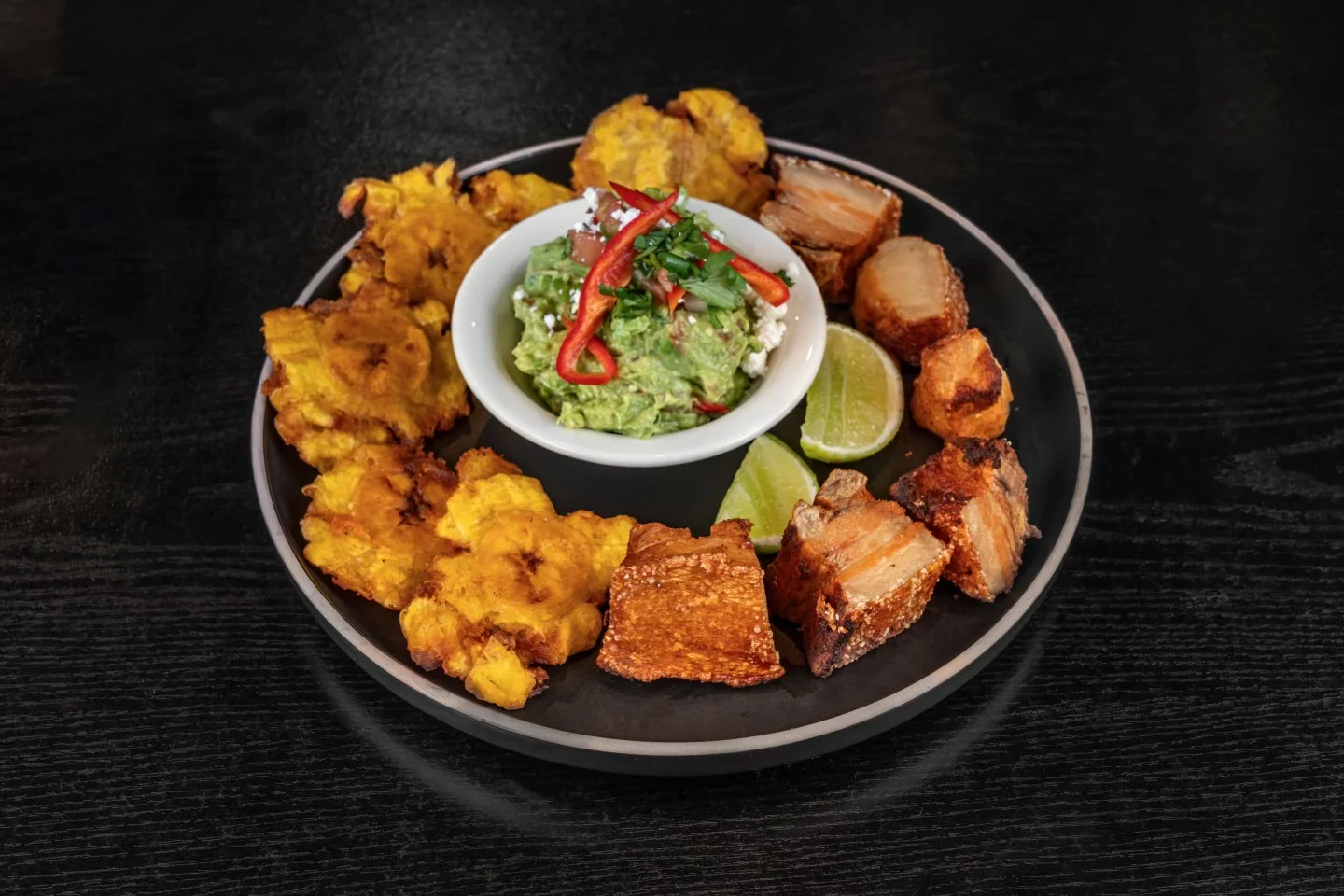 A platter featuring fried plantains, crispy pork, and a bowl of guacamole garnished with fresh herbs.