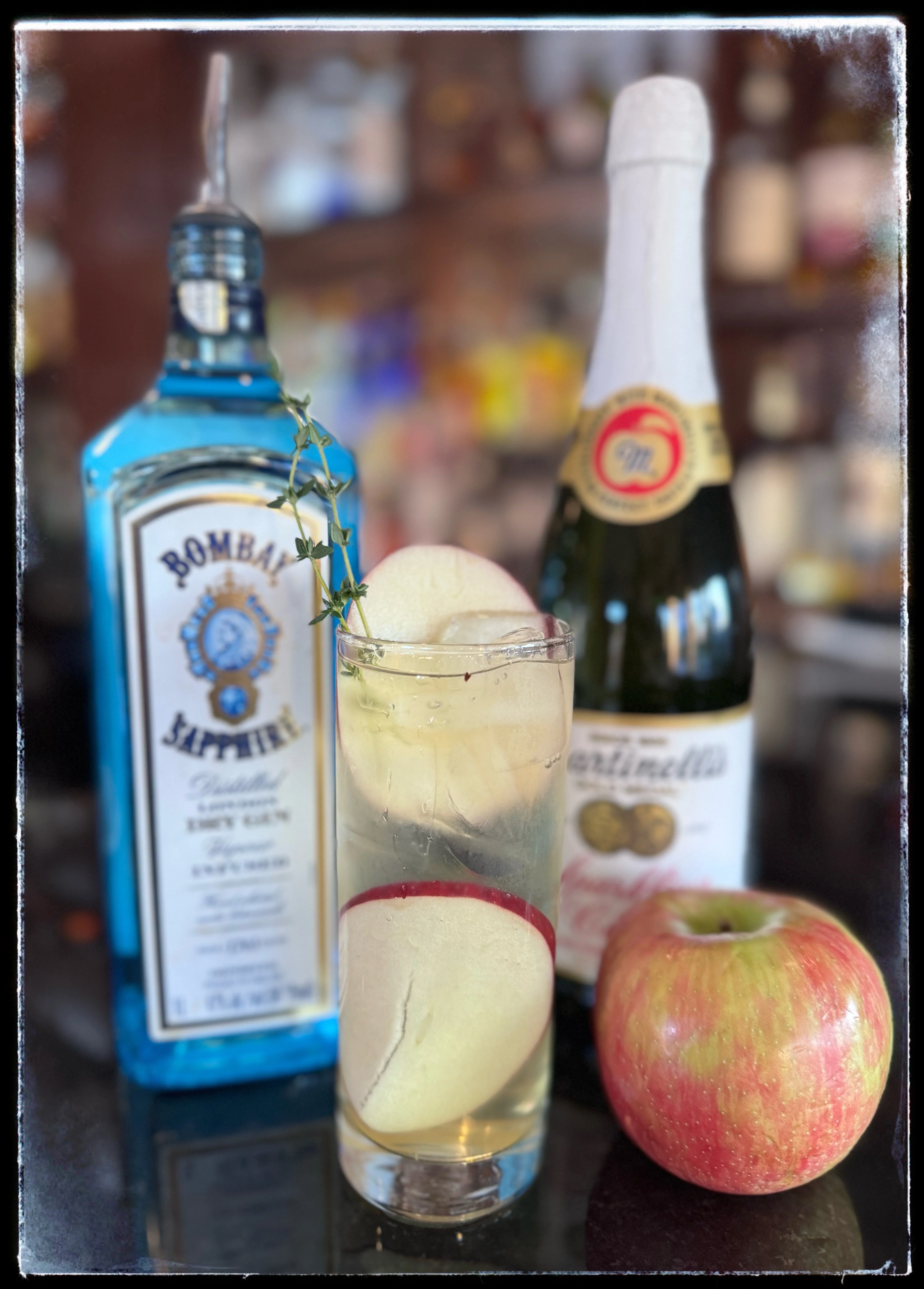 A cocktail garnished with apple slices and thyme is in front of gin and sparkling cider bottles. A fresh apple sits beside the glass on a bar counter.