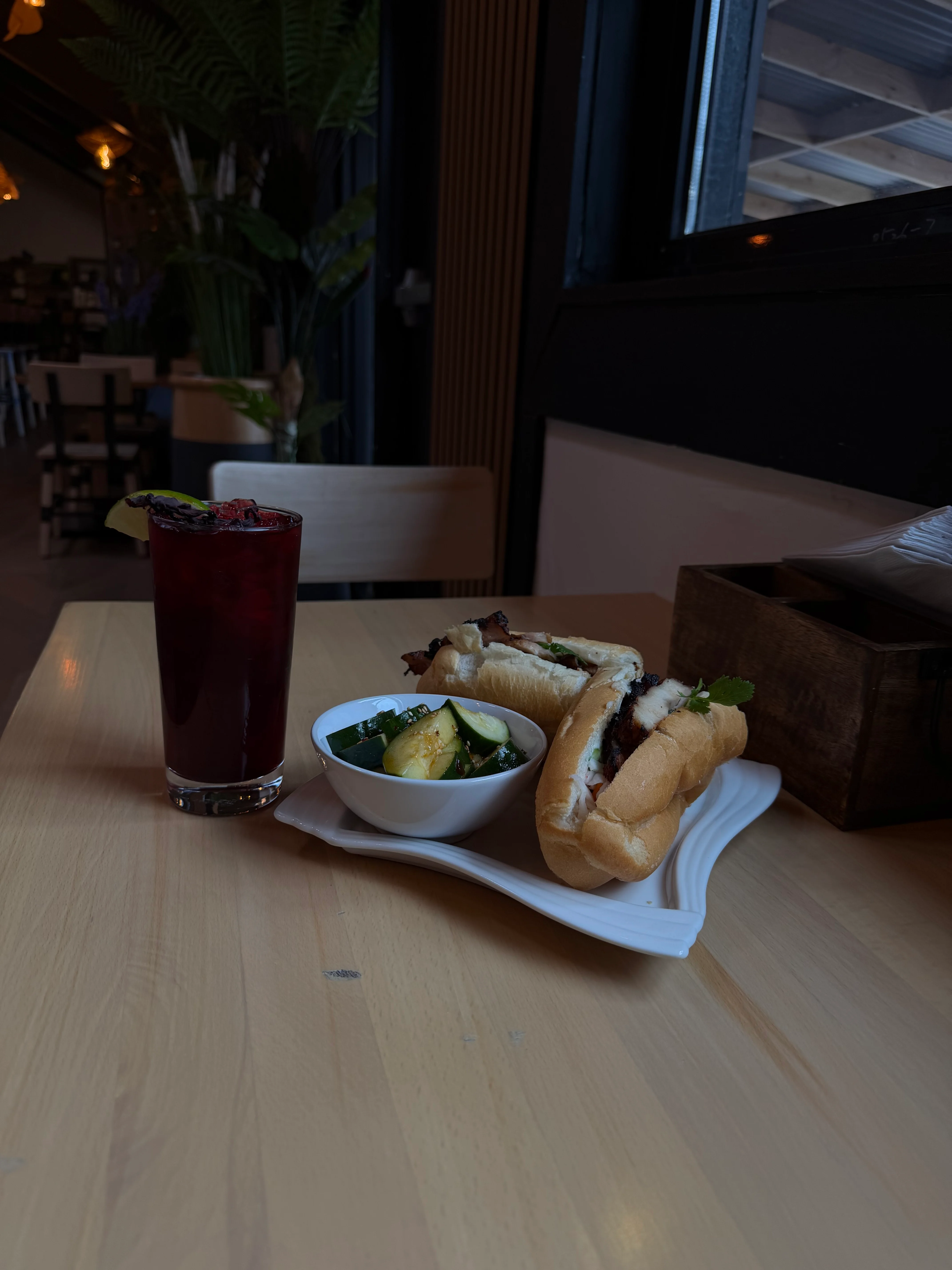 A dish featuring two hot dogs topped with garnishes, accompanied by a refreshing drink and a small bowl of pickled vegetables. The setting is a cozy dining area.