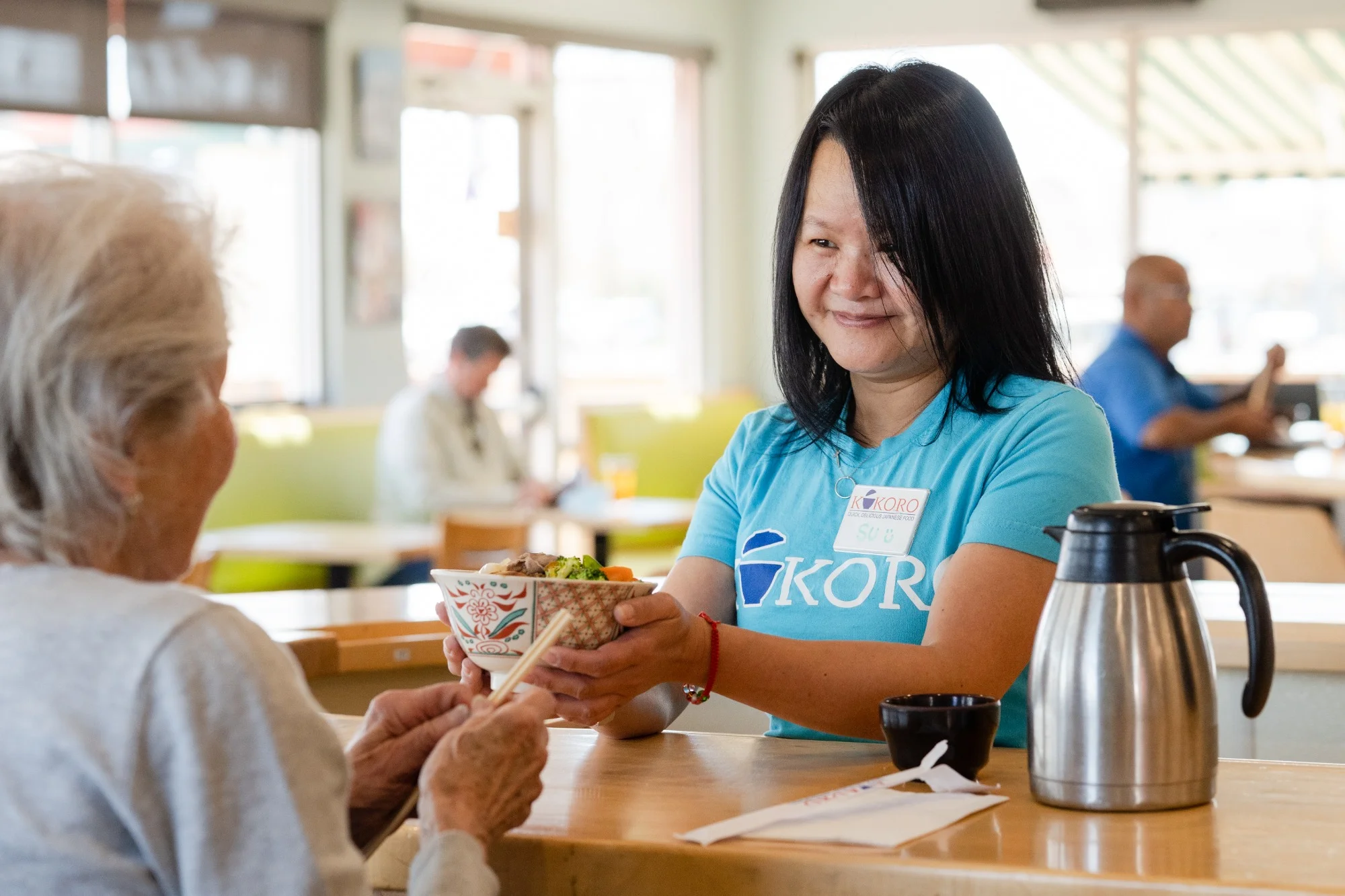 A restaurant scene where a server hands a bowl of food to an elderly customer, with others dining in the background.
