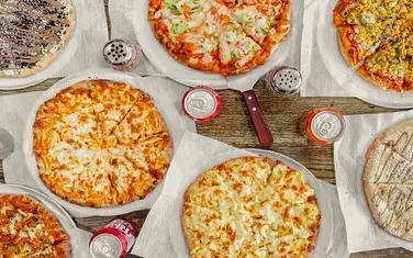 An assortment of pizzas displayed on a table, featuring various toppings and styles, accompanied by beverages and seasonings.