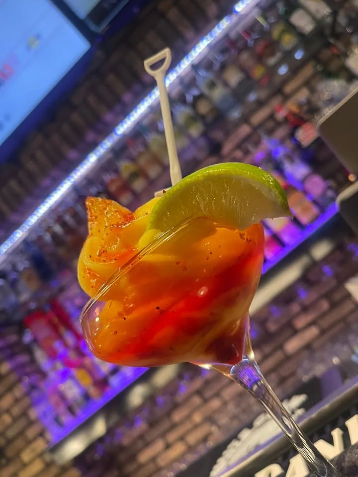 A colorful cocktail with layers of orange, yellow, and red, garnished with lime and shrimp on a skewer, served in a glass.